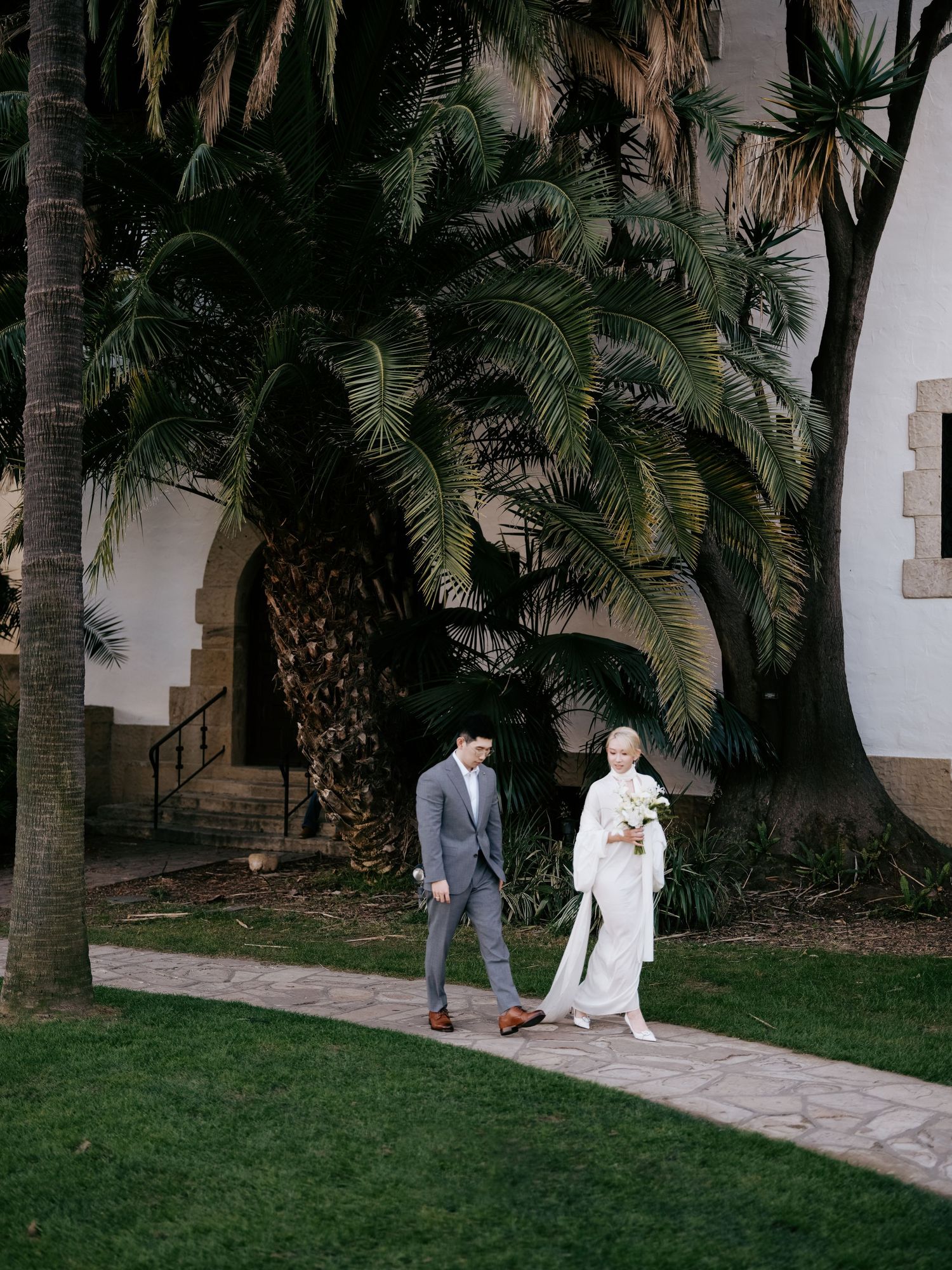 Bride and groom stroll along garden path beneath towering palm trees at venue.