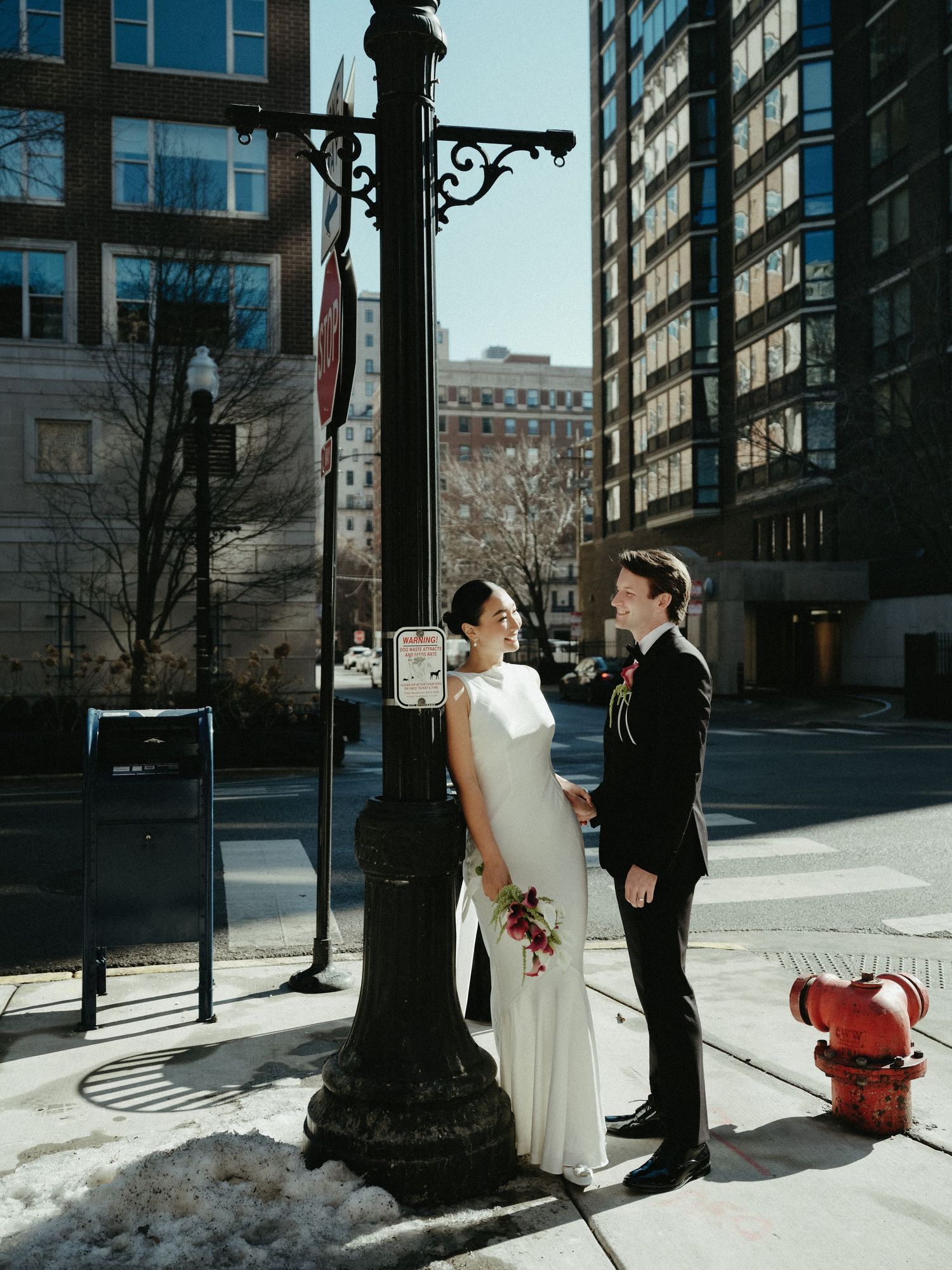 Bride and groom share a joyful moment beside a vintage street lamp on a sunny urban city sidewalk.