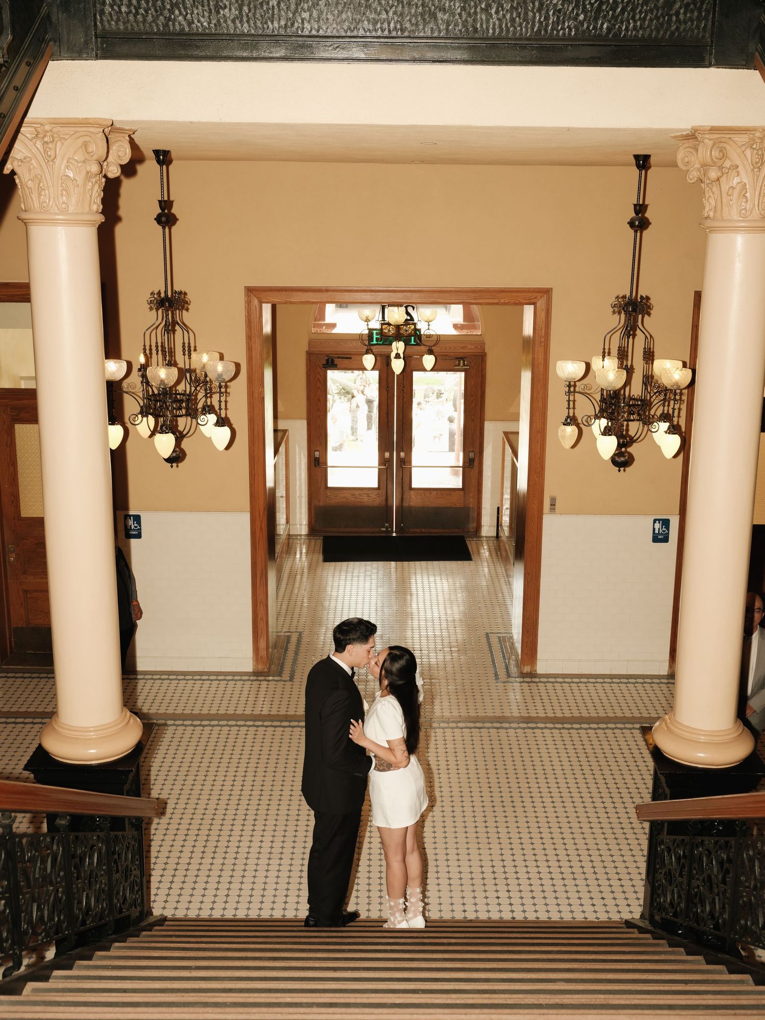 Newlyweds share a kiss beneath grand chandeliers in ornate tiled hallway with decorative columns.