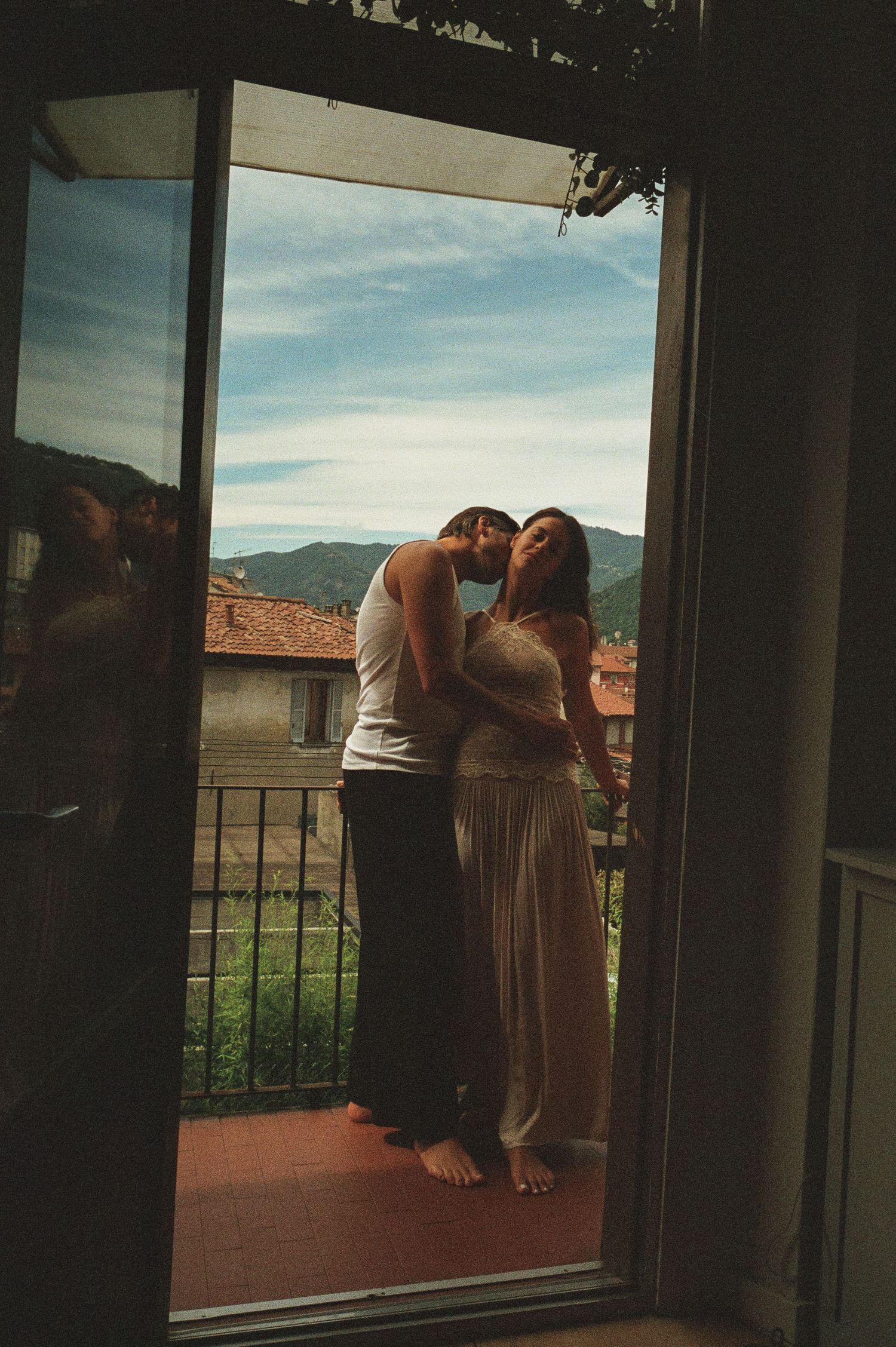 Couple embracing intimately on a balcony doorway with lush mountain landscape in background.
