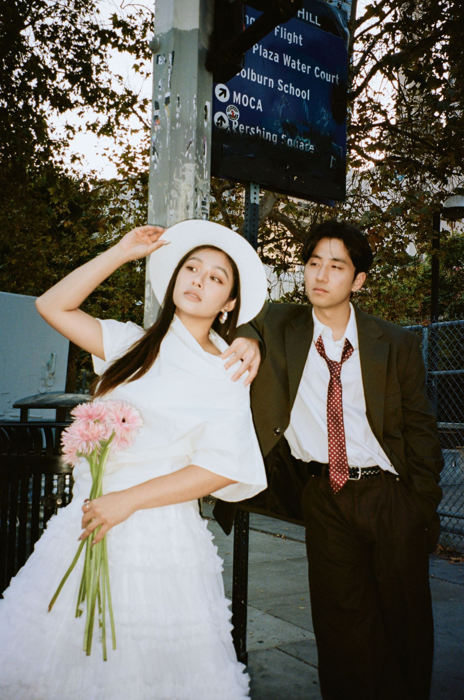 Stylish bride in white dress and hat holds pink flowers beside suited groom near MOCA street sign in LA.