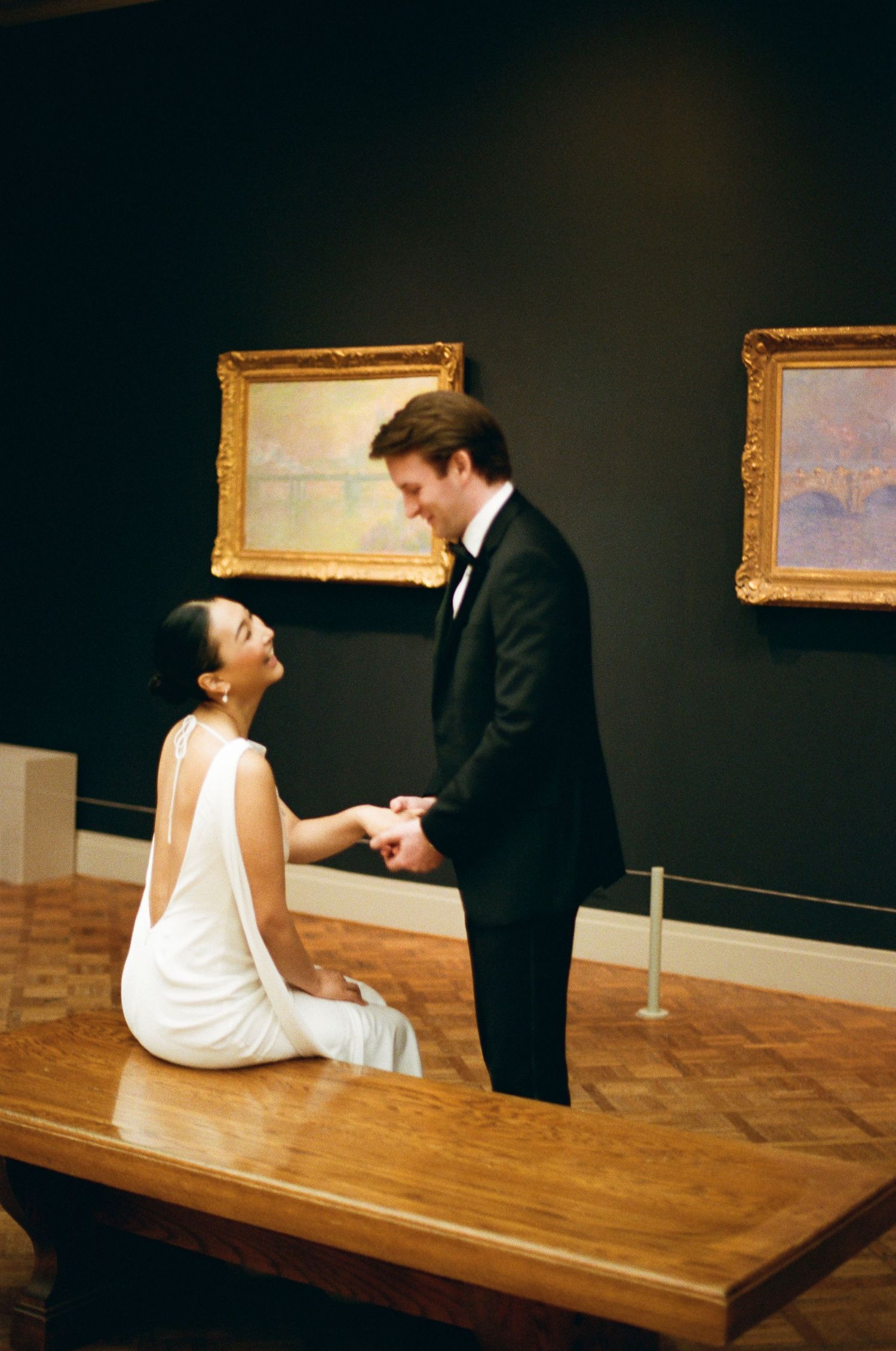 Bride sits on a museum bench holding the groom's hands, surrounded by Impressionist paintings in golden frames.