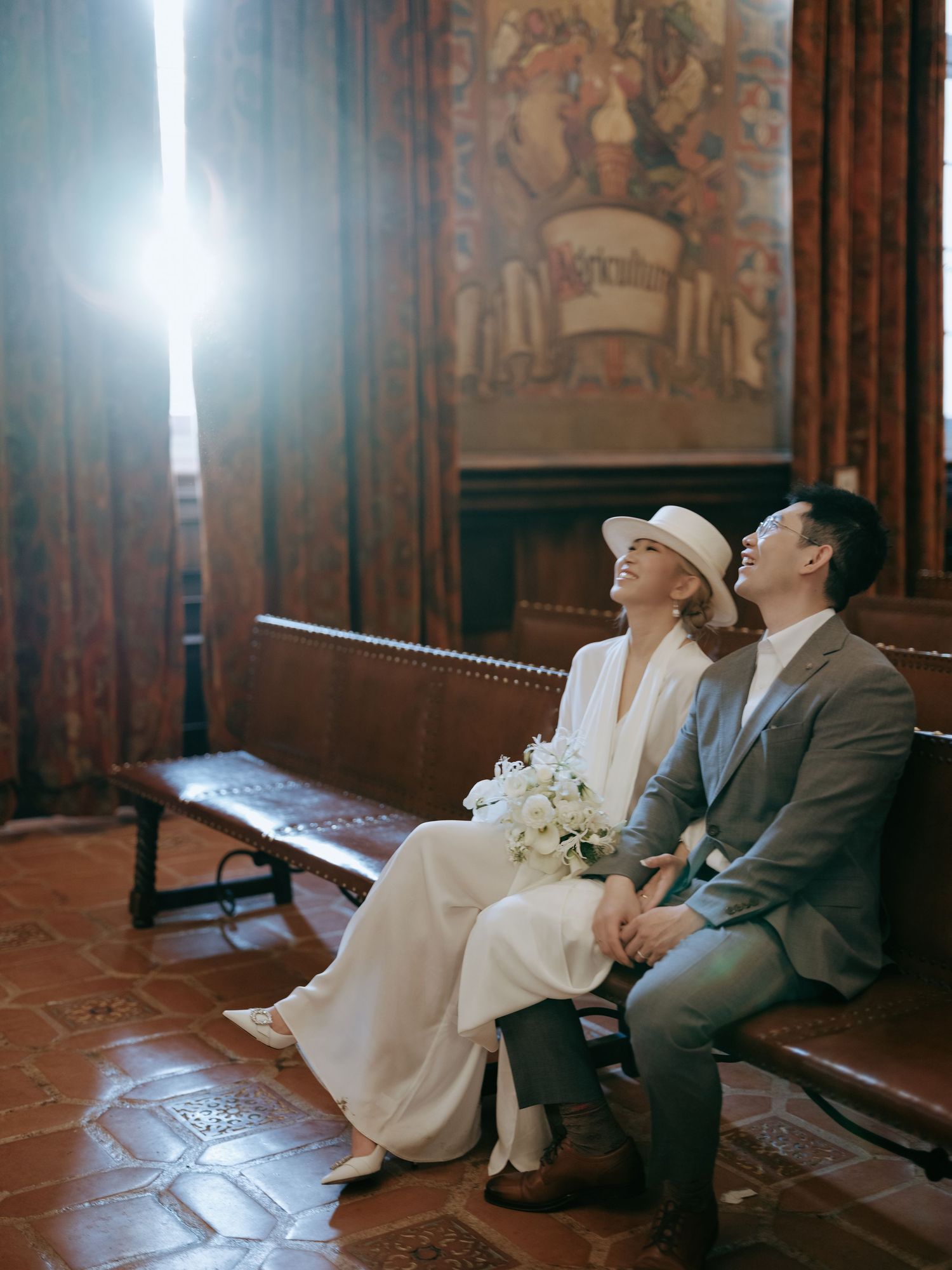Bride and groom sitting on wooden bench in historic room admiring ornate wall murals together.