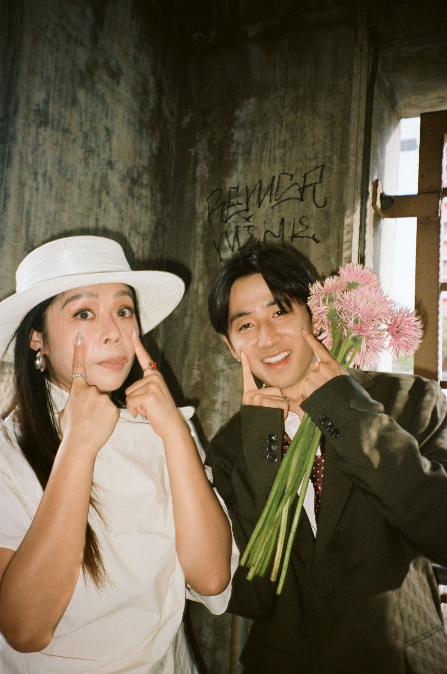 Smiling couple makes cute face gestures indoors, woman in white hat, man in dark suit holding pink gerbera flowers.