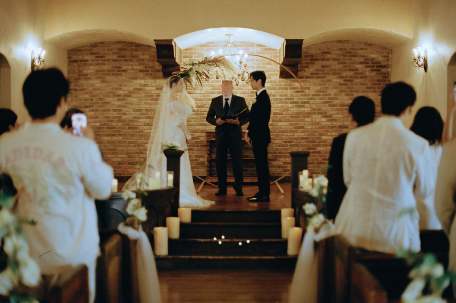 Bride and groom exchange vows at candlelit altar in intimate brick-walled wedding chapel.