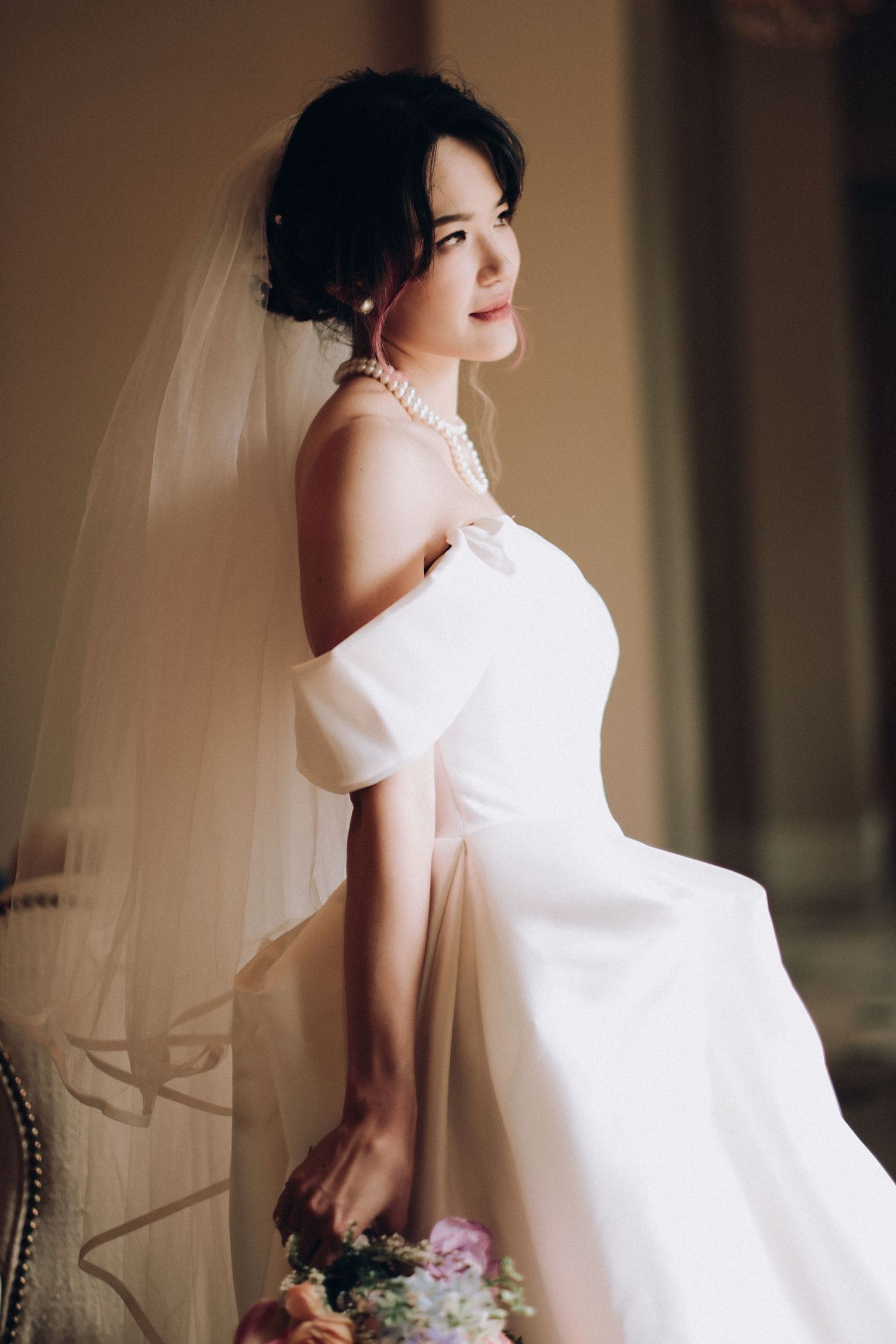 Elegant bride in off-shoulder white gown with veil and pearl necklace poses in soft indoor light.