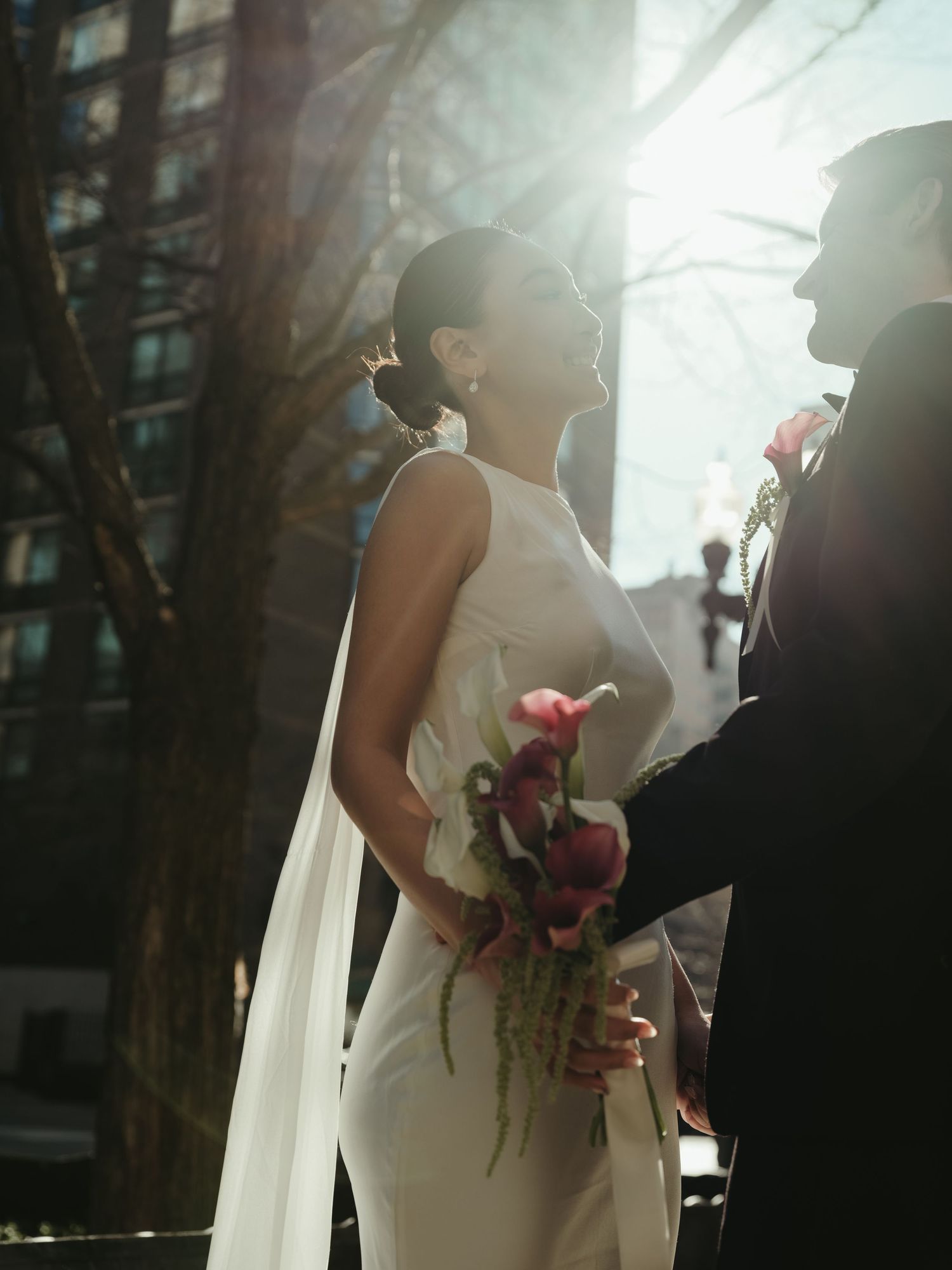 Bride in white gown holds pink calla lily bouquet as sunlight halos couple during outdoor wedding portraits.
