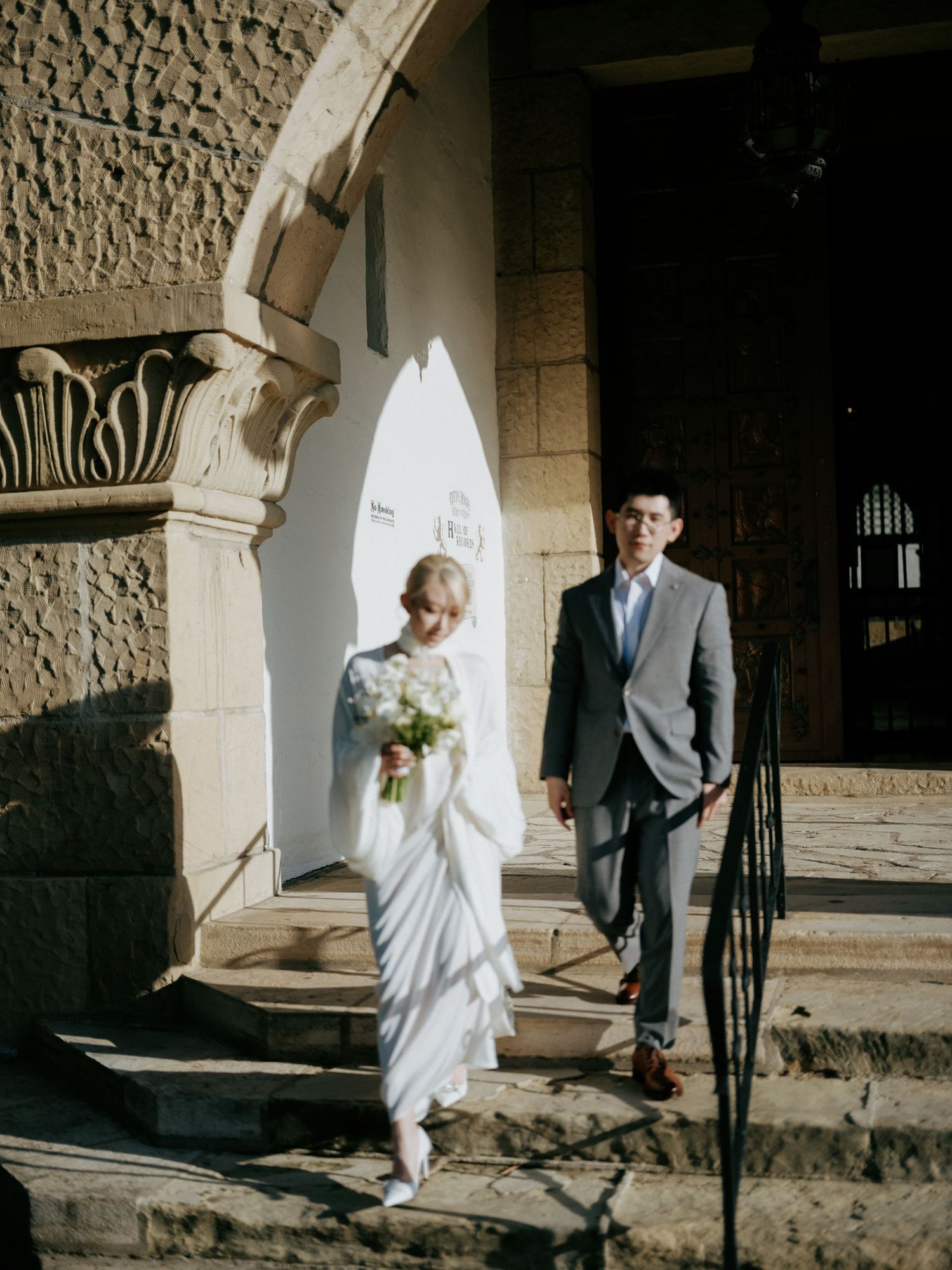 Bride carrying white floral bouquet walks with groom under stone arch in sunlight.