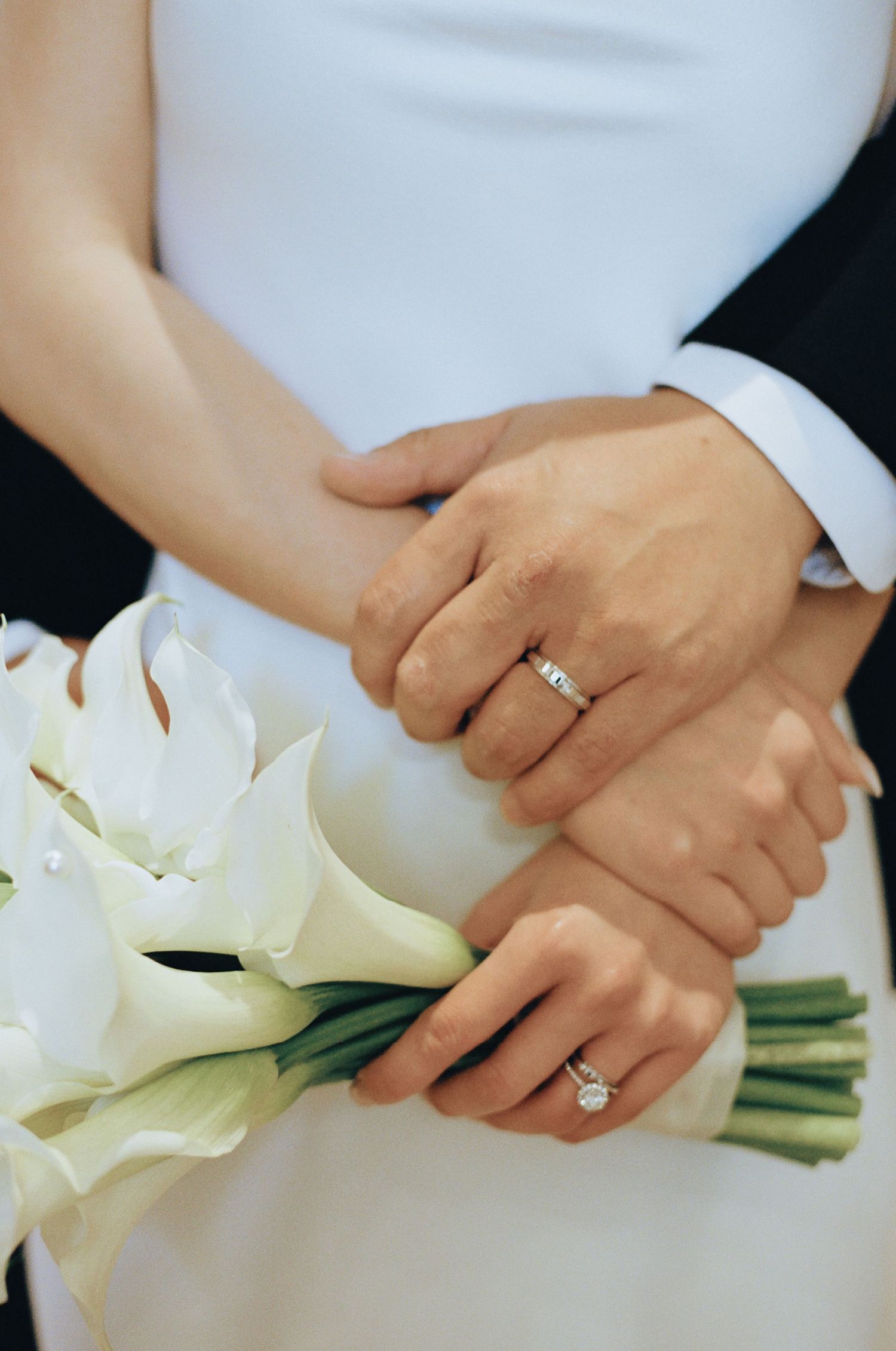 Bride and groom holding white calla lily bouquet, showcasing elegant wedding bands on their hands.