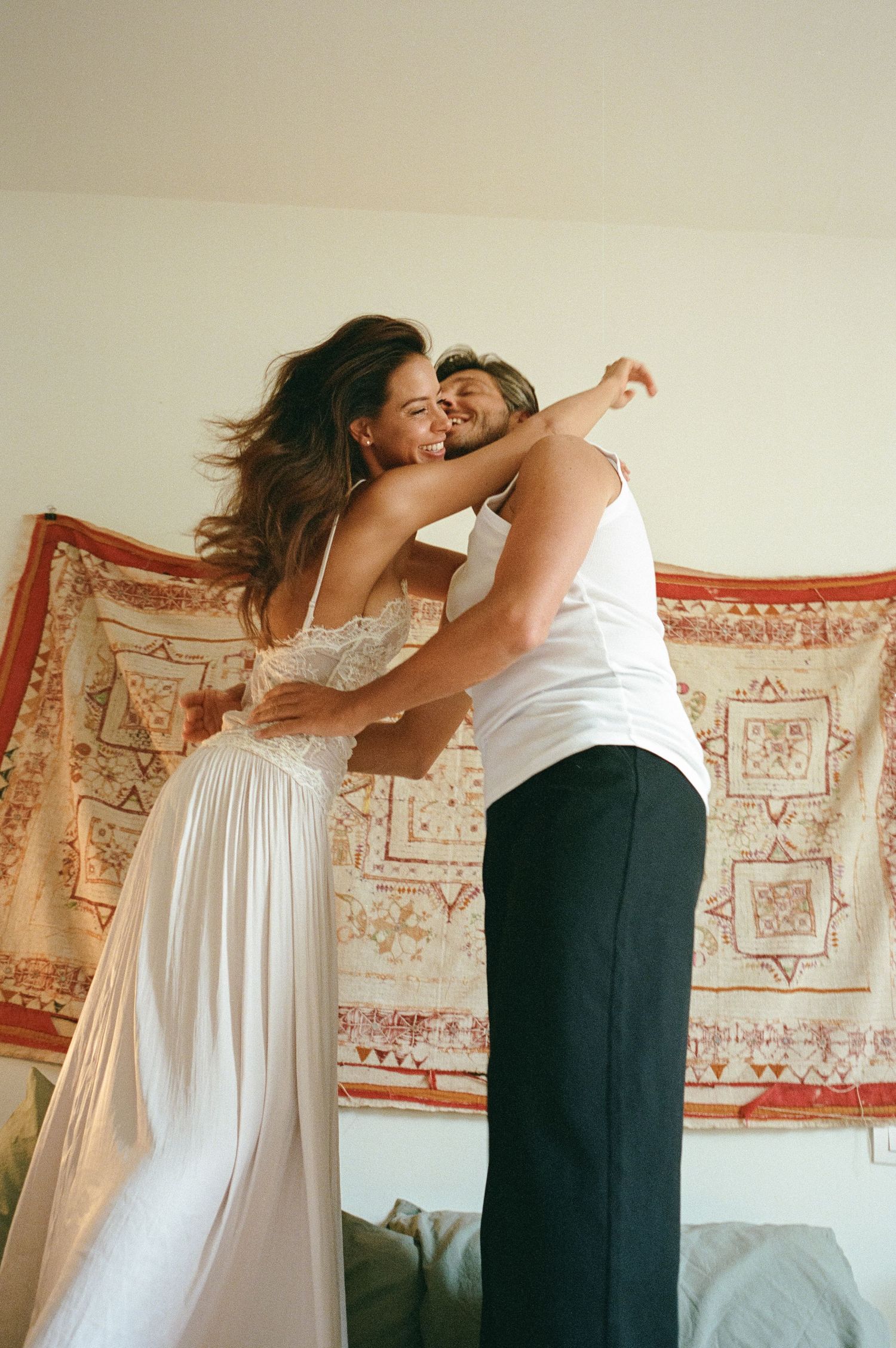 Joyful couple dancing and hugging in front of a bohemian patterned tapestry indoors.