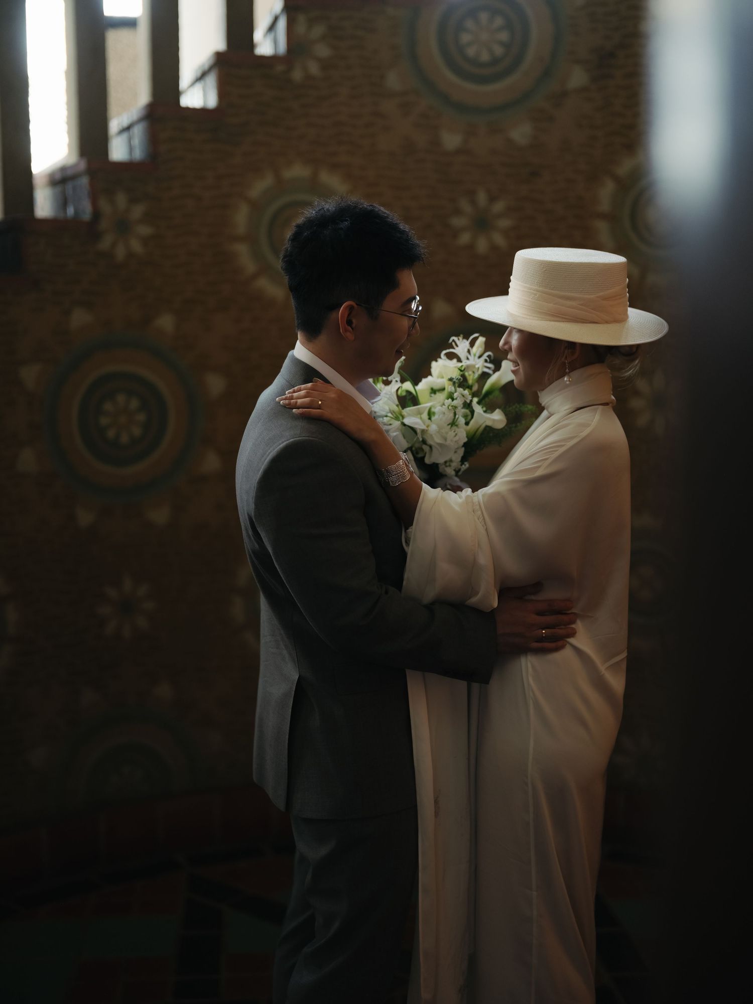 Bride and groom embrace tenderly, she holds white floral bouquet against decorative tiled wall.