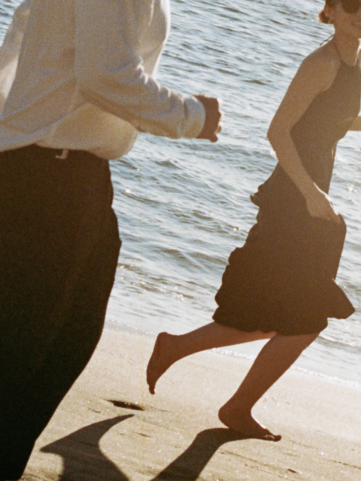 Barefoot couple running together along sunny beach shoreline near ocean waves.