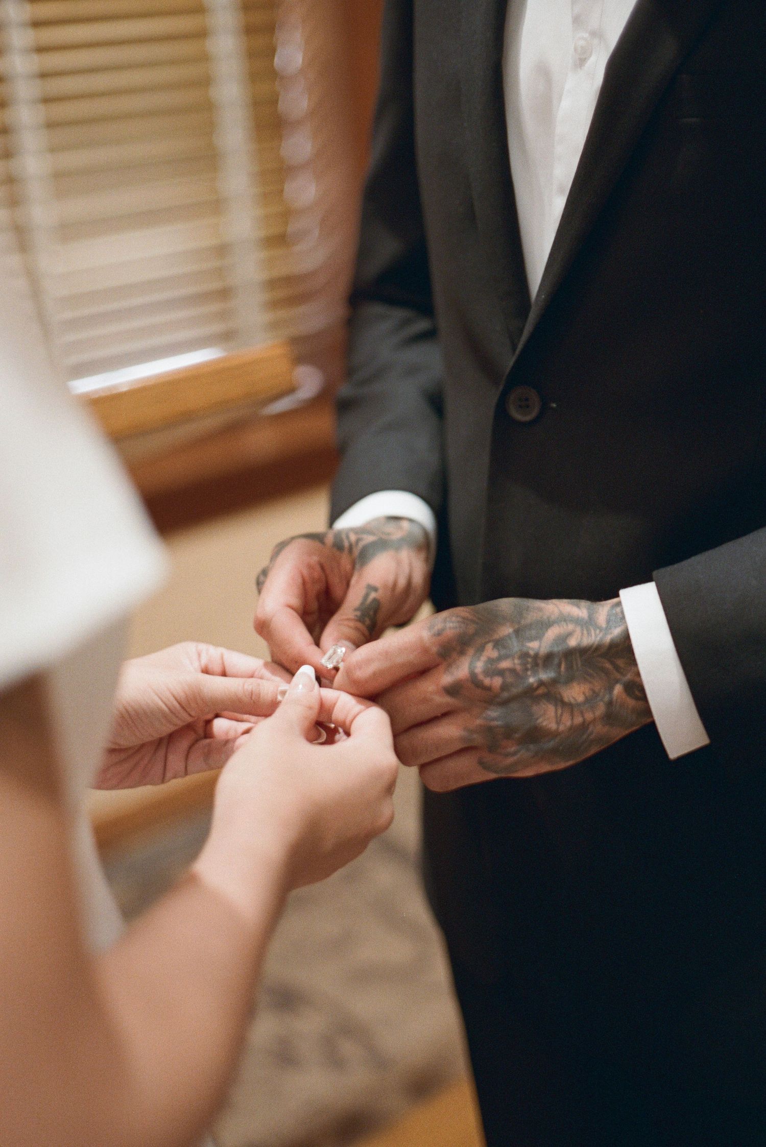 Groom with tattooed hands placing a wedding ring on bride's finger during intimate ceremony.
