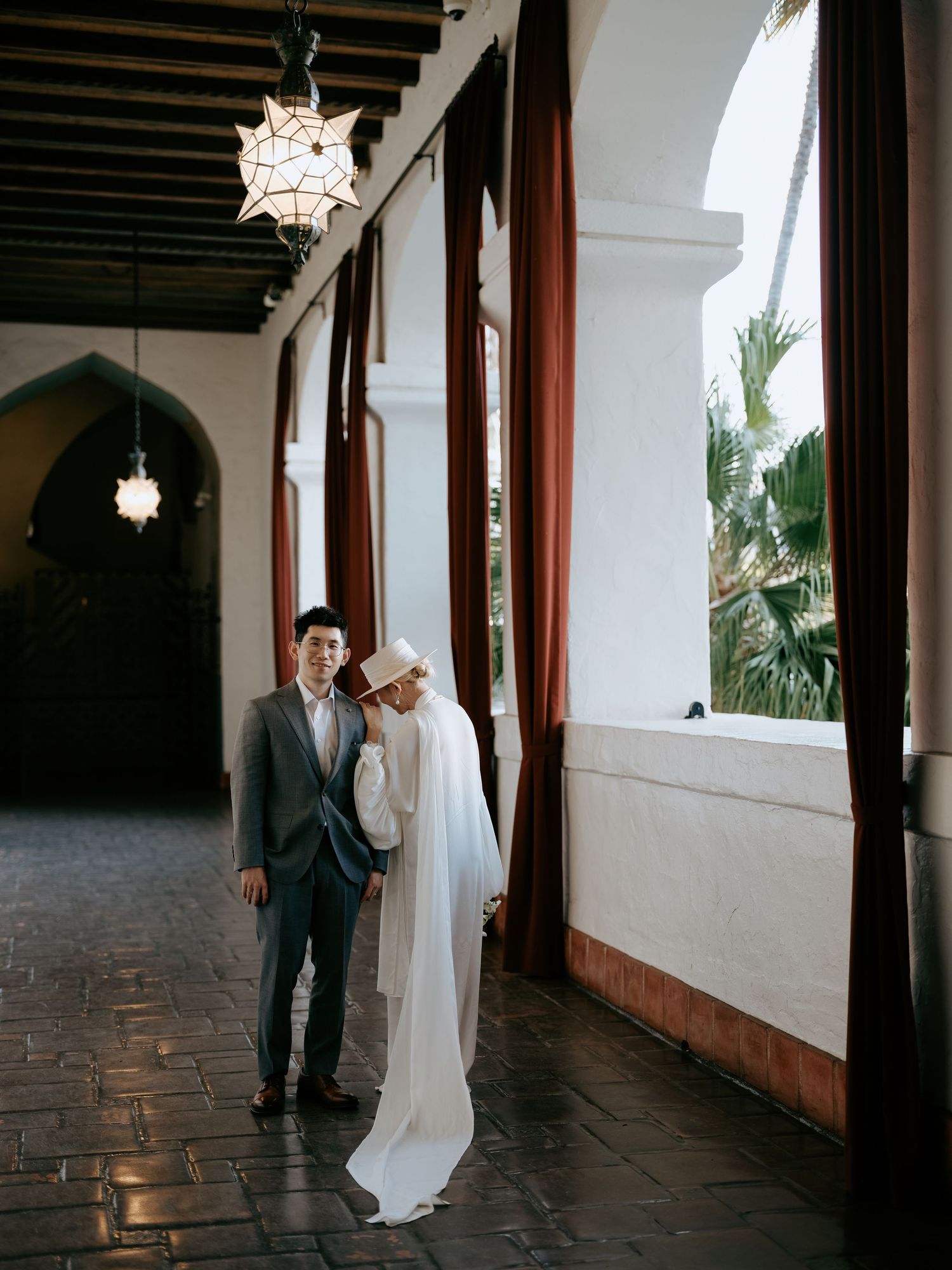 Bride in flowing white dress and hat leans on groom in gray suit in sunlit arched hallway.
