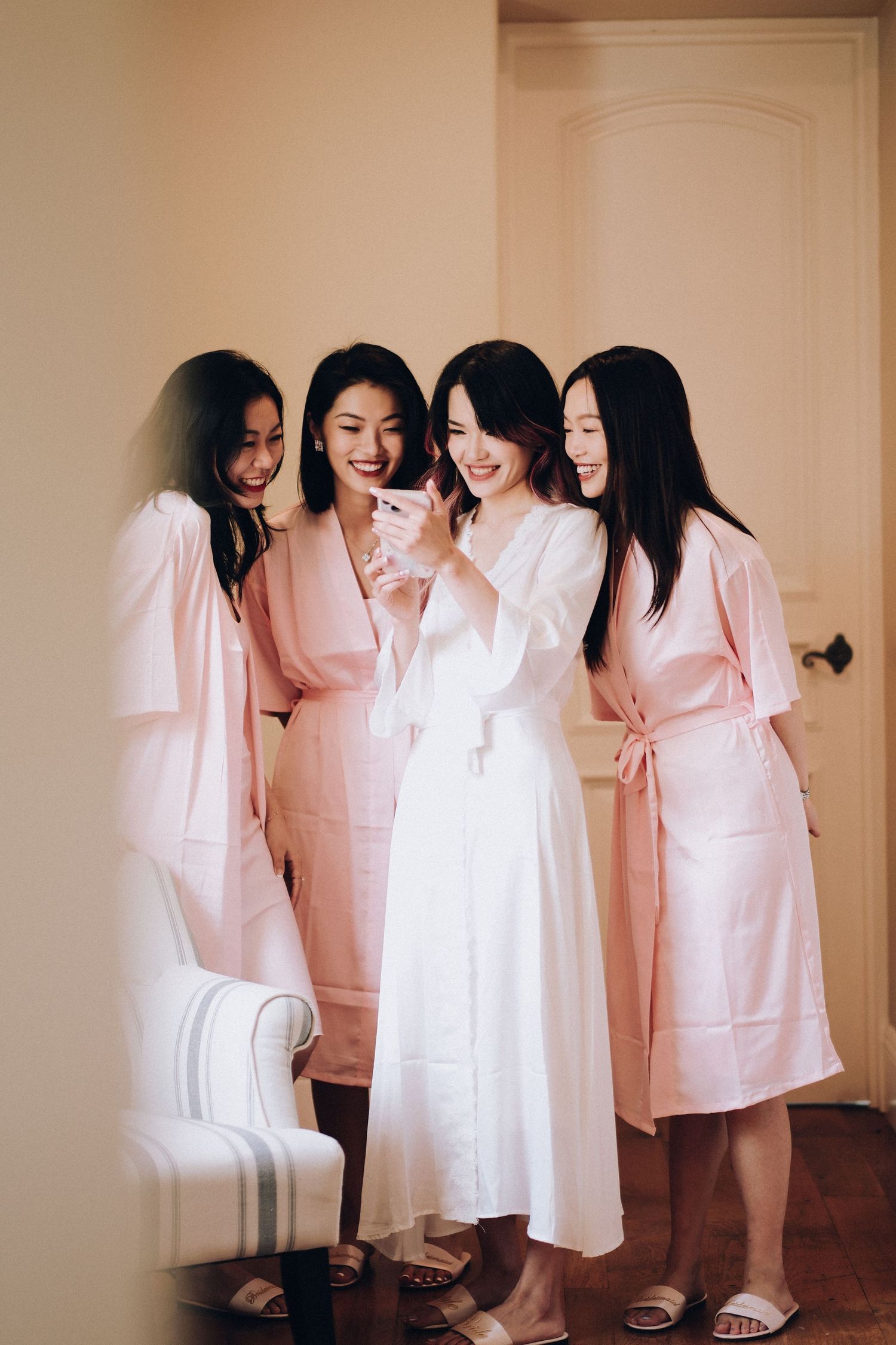 Bride in white robe laughing with three bridesmaids in pink robes during wedding preparations.