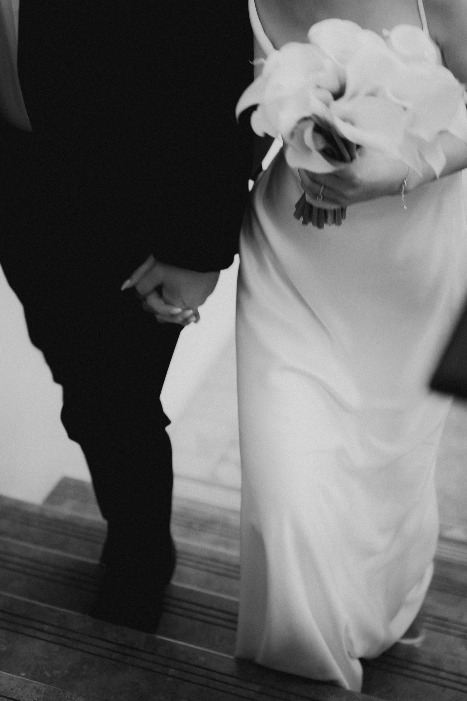 Black and white photo of bride and groom holding hands, bride carrying white floral bouquet in elegant satin gown.