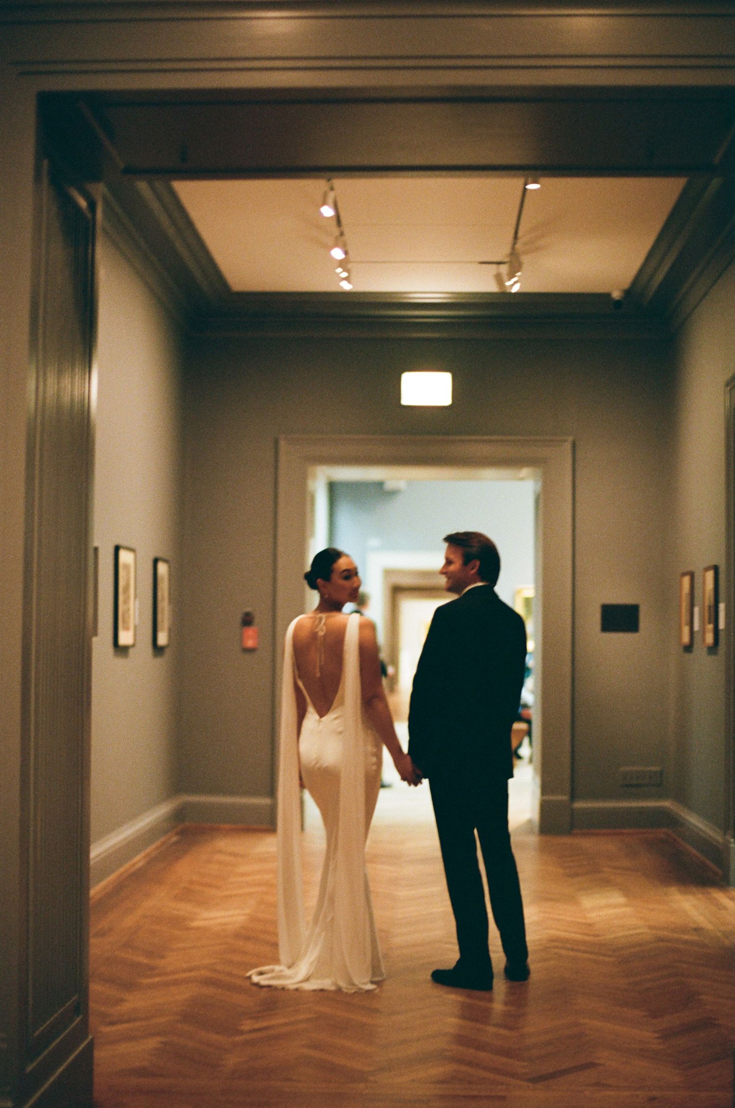 Bride and groom hold hands in a softly lit museum hallway, the bride's open back gown on full display.