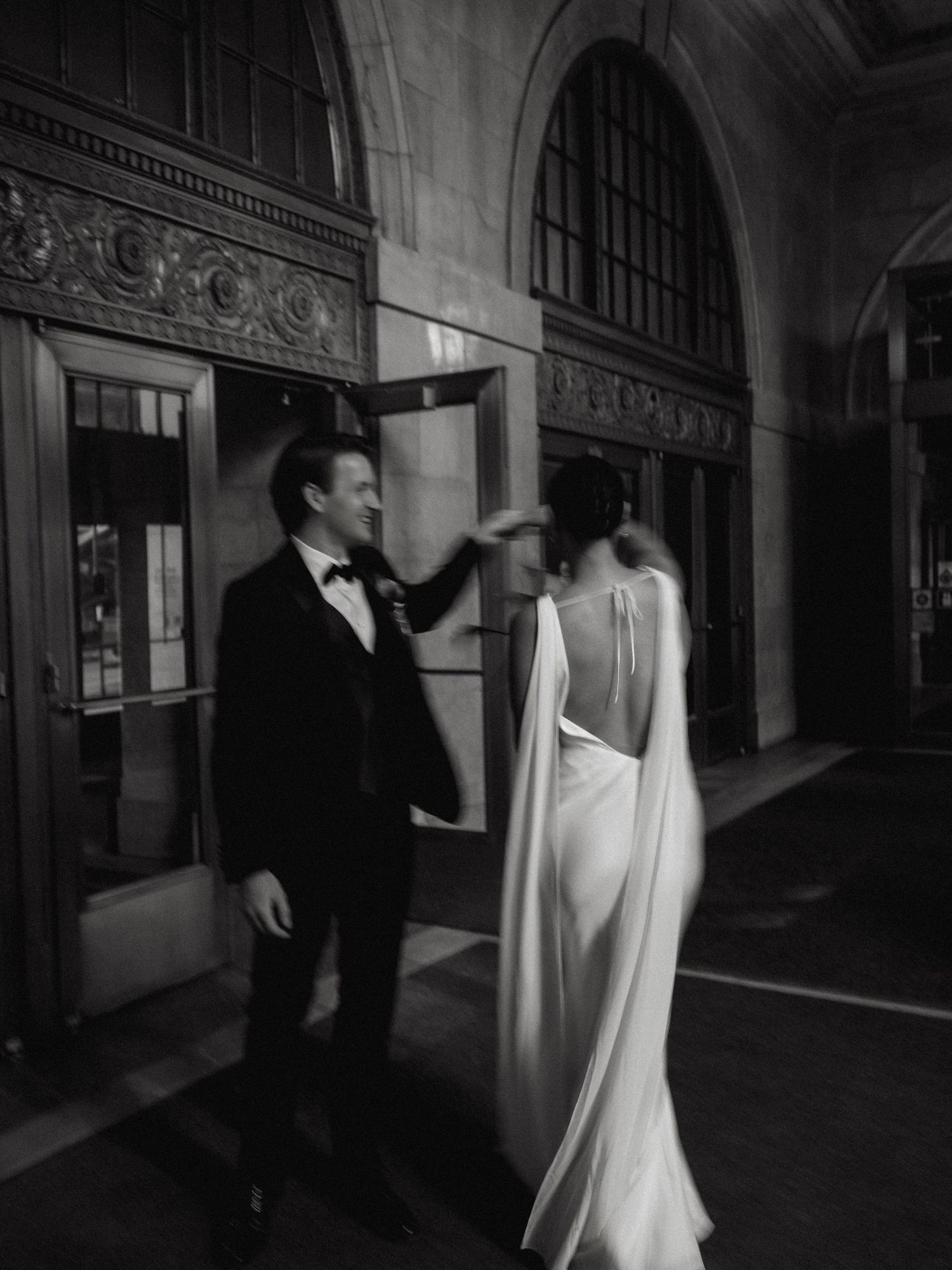 Black and white photo of groom adjusting brides flowing cape gown outside elegant venue entrance.