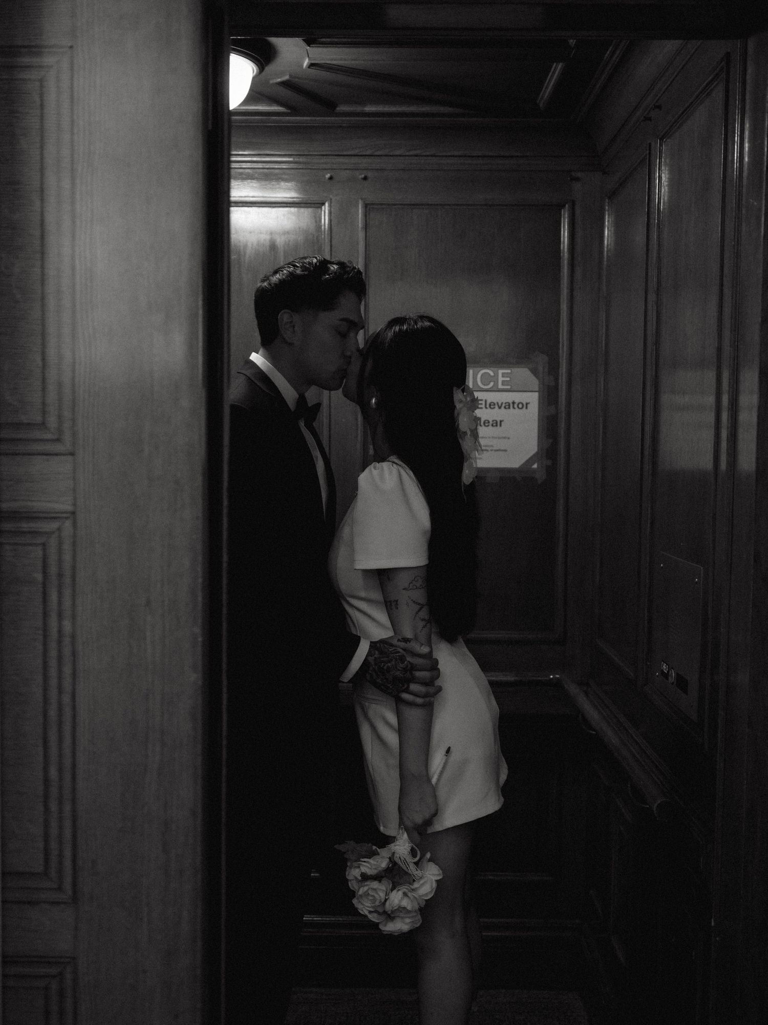 Newlyweds share a kiss in black and white photo near an elevator door, bride holding bouquet.