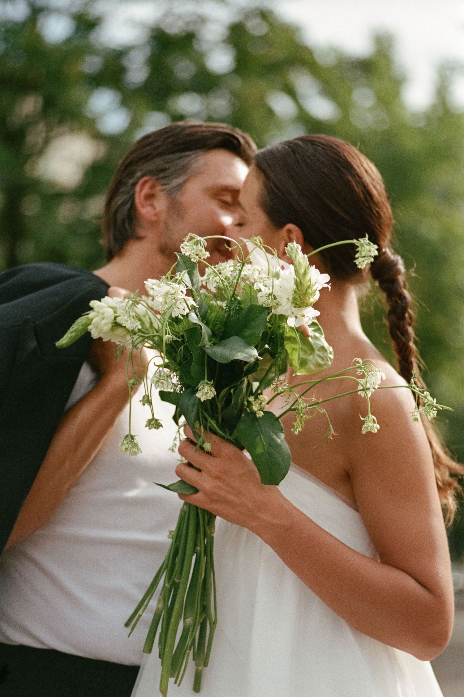 Bride and groom kissing while holding a beautiful white wildflower bouquet in an outdoor setting.