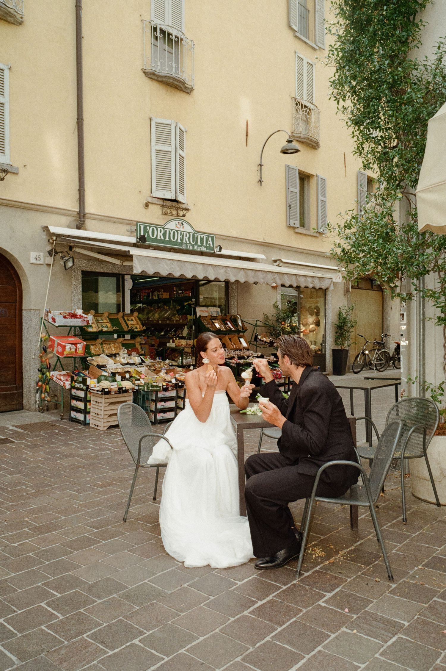 Bride and groom sharing drinks at an outdoor cafe table in front of an Italian ortofrutta market.