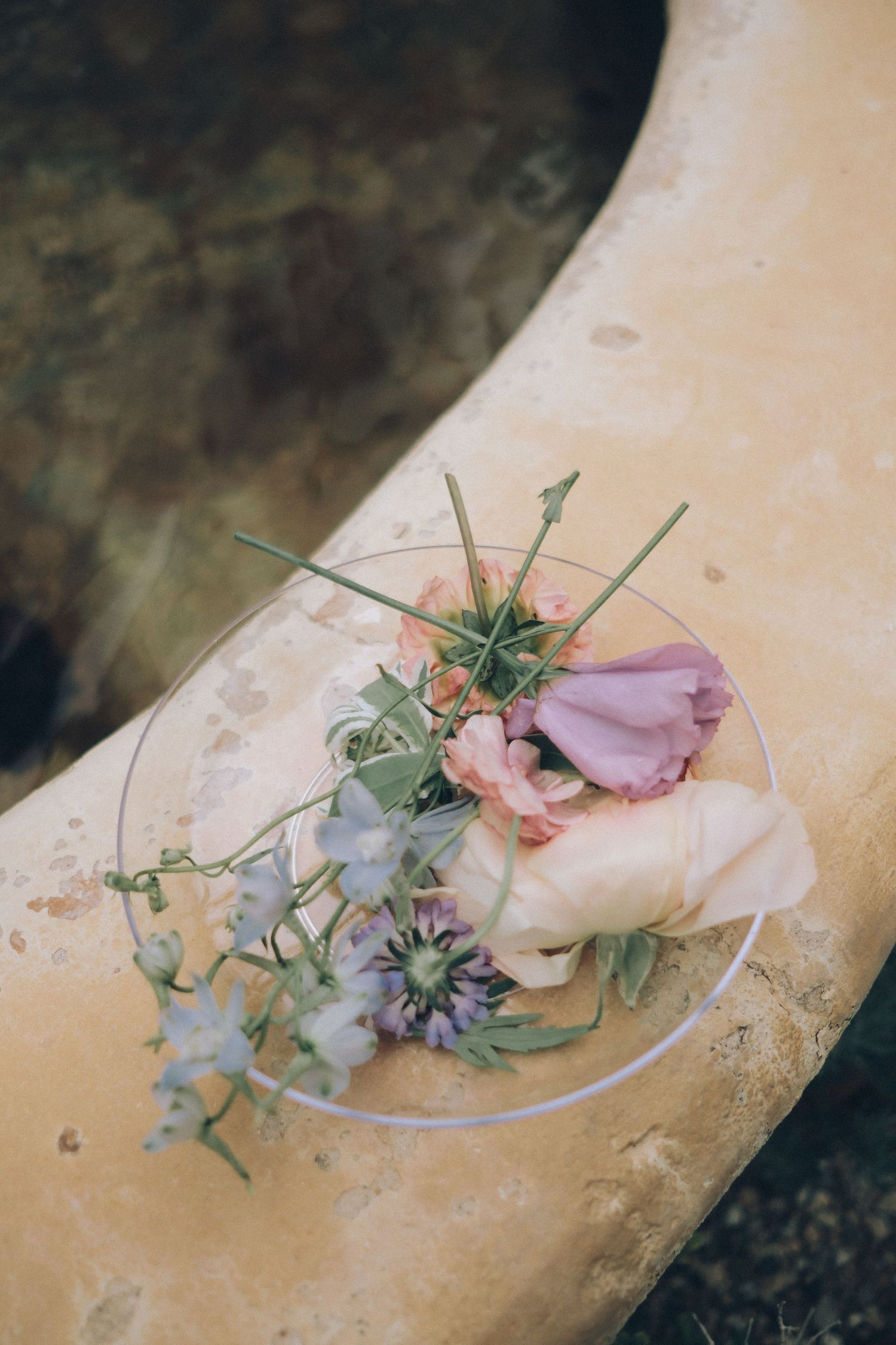 Delicate floral arrangement with pastel petals and greenery resting on stone fountain edge.