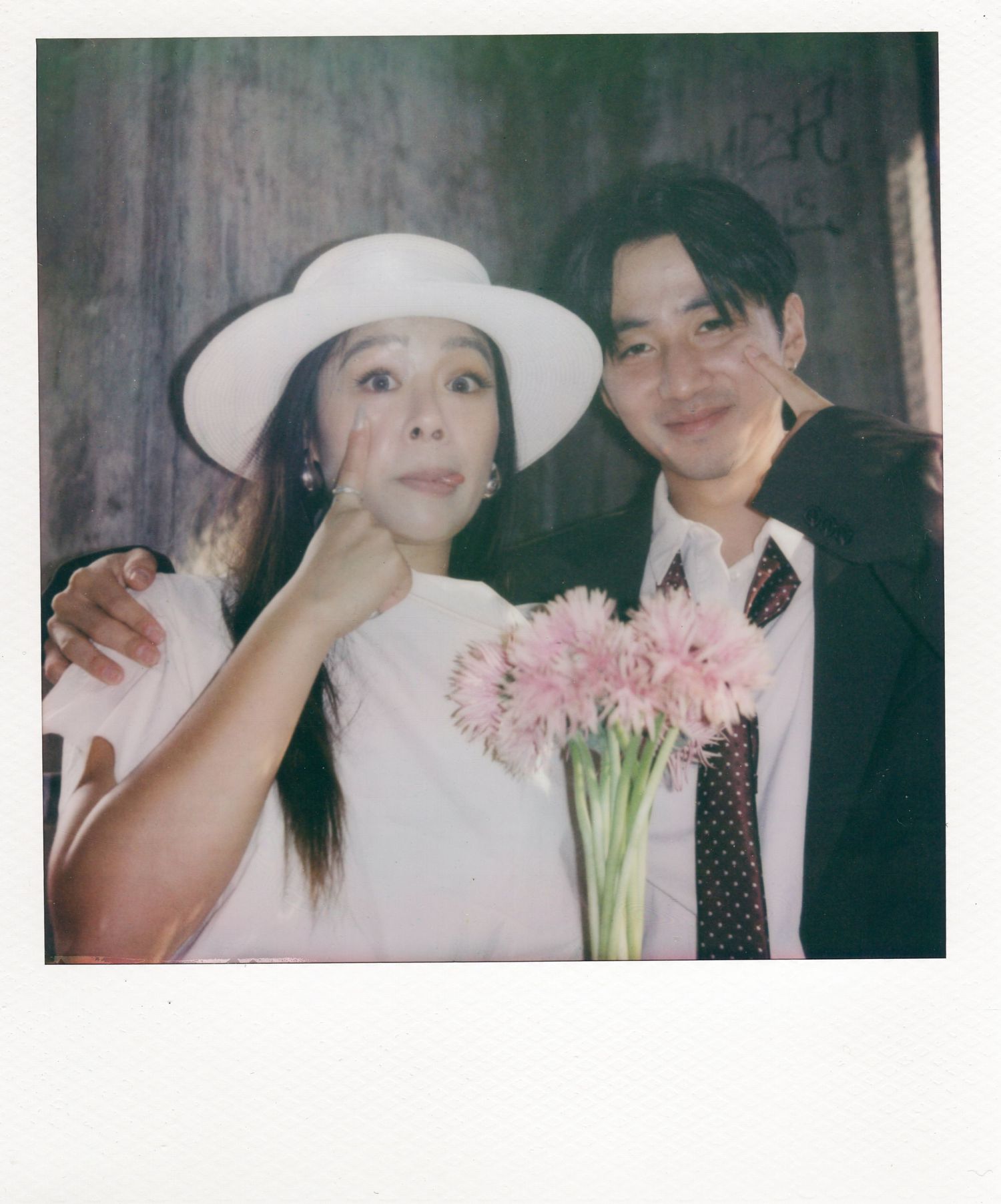 Smiling couple poses together, bride in white hat holds pink flowers in a vintage Polaroid-style portrait.