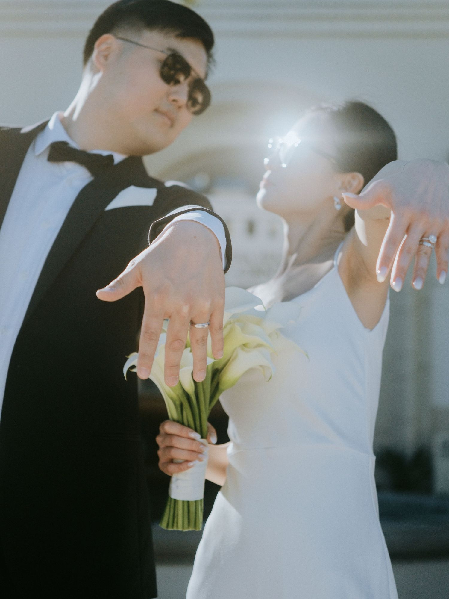 Wedding couple showing off rings with white bouquet in bright sunlight outdoors.