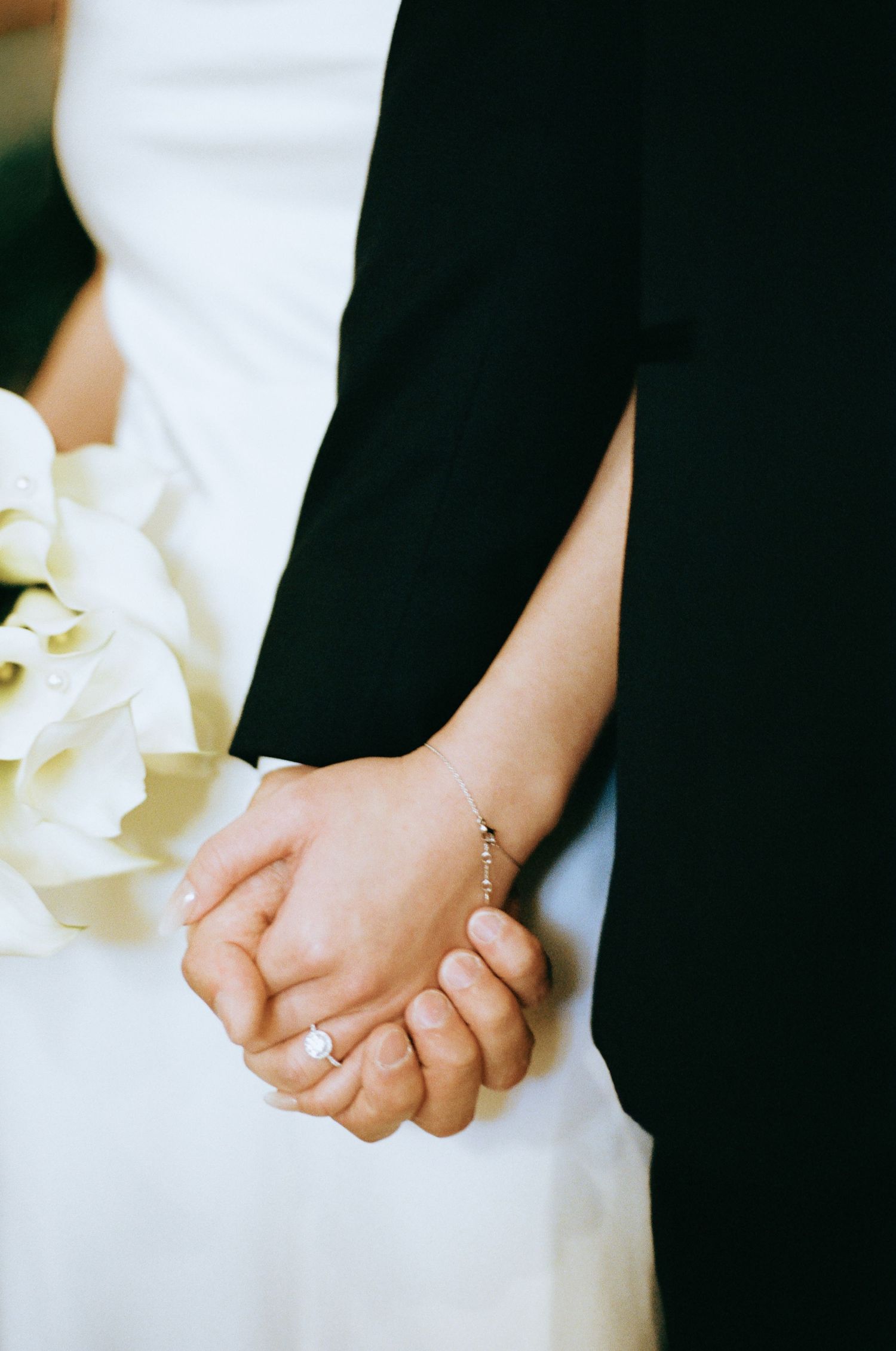 Close-up of bride and groom holding hands, bride wearing engagement ring and holding white floral bouquet.