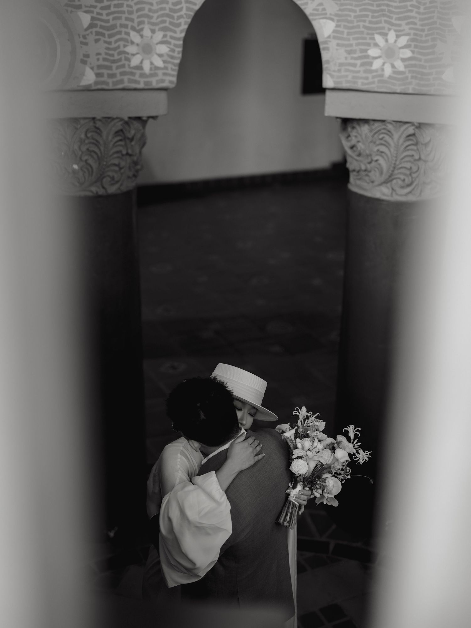 Black and white photo of bride in wide-brim hat embracing groom while holding floral bouquet.