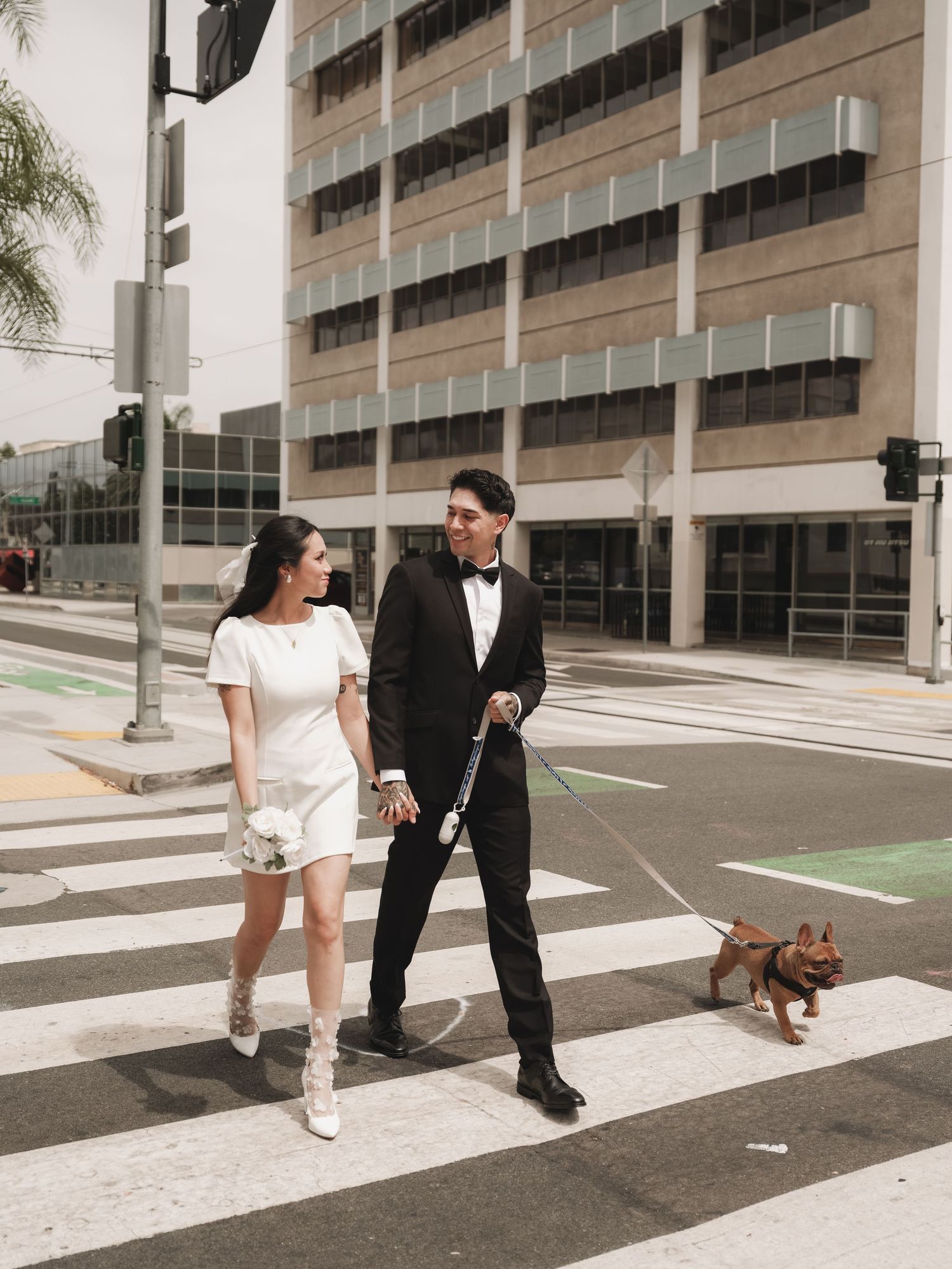 Smiling newlywed couple walking French Bulldog across city crosswalk in wedding attire.
