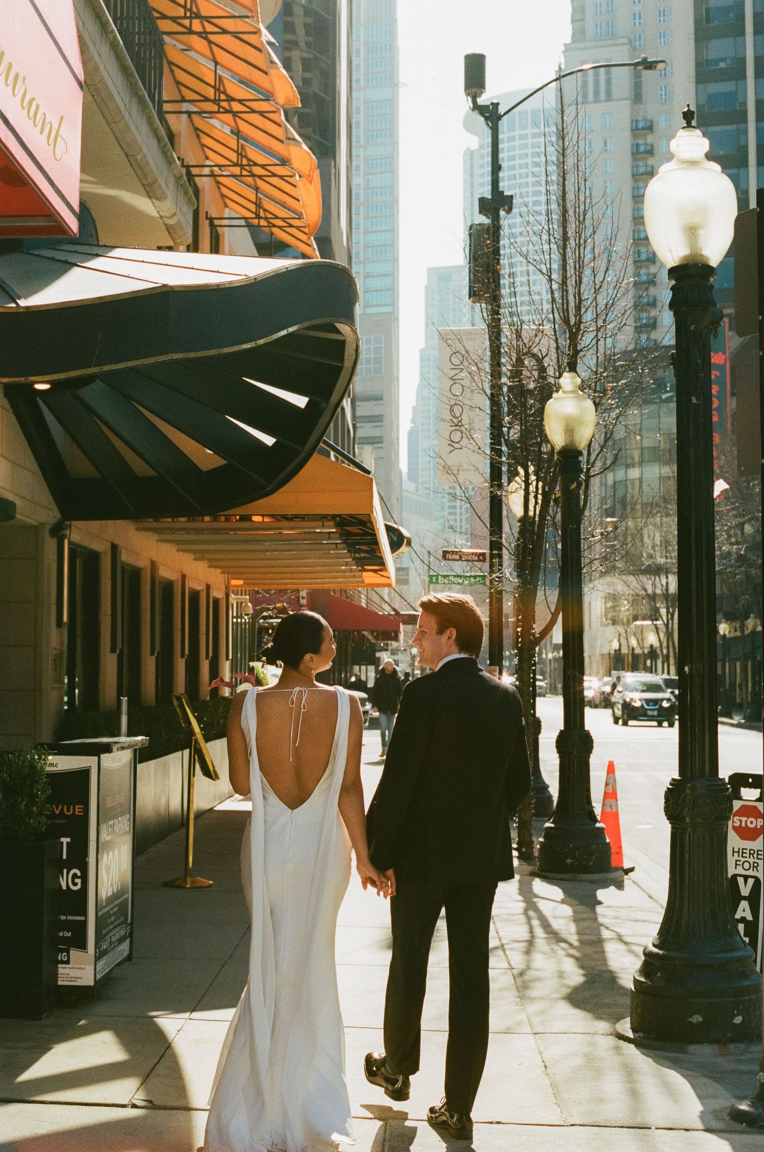 Bride and groom walk hand in hand down sunny Chicago sidewalk past ornate theater awning.