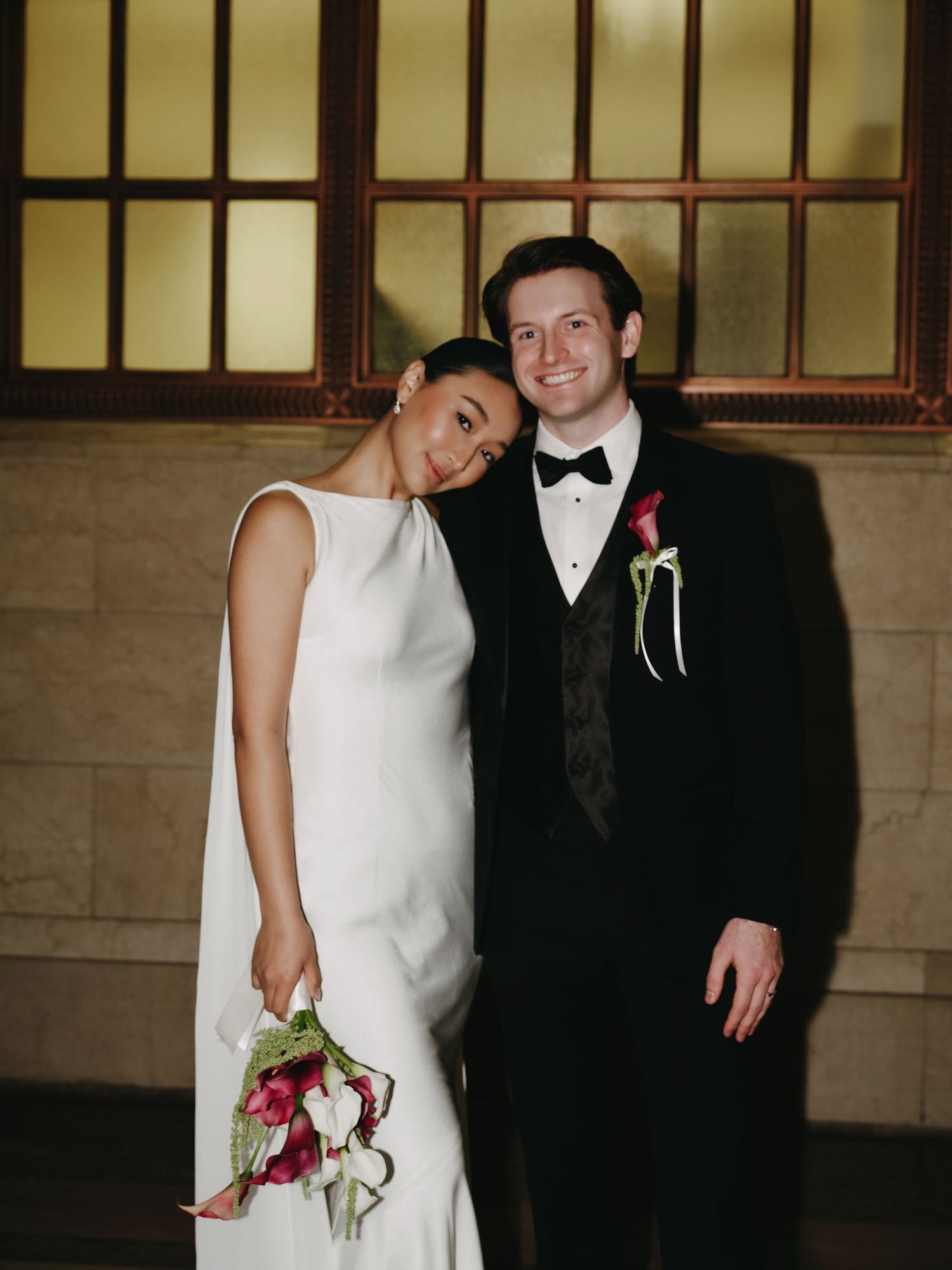 Bride and groom posing together smiling in ornate indoor venue, bride holding calla lily bouquet.