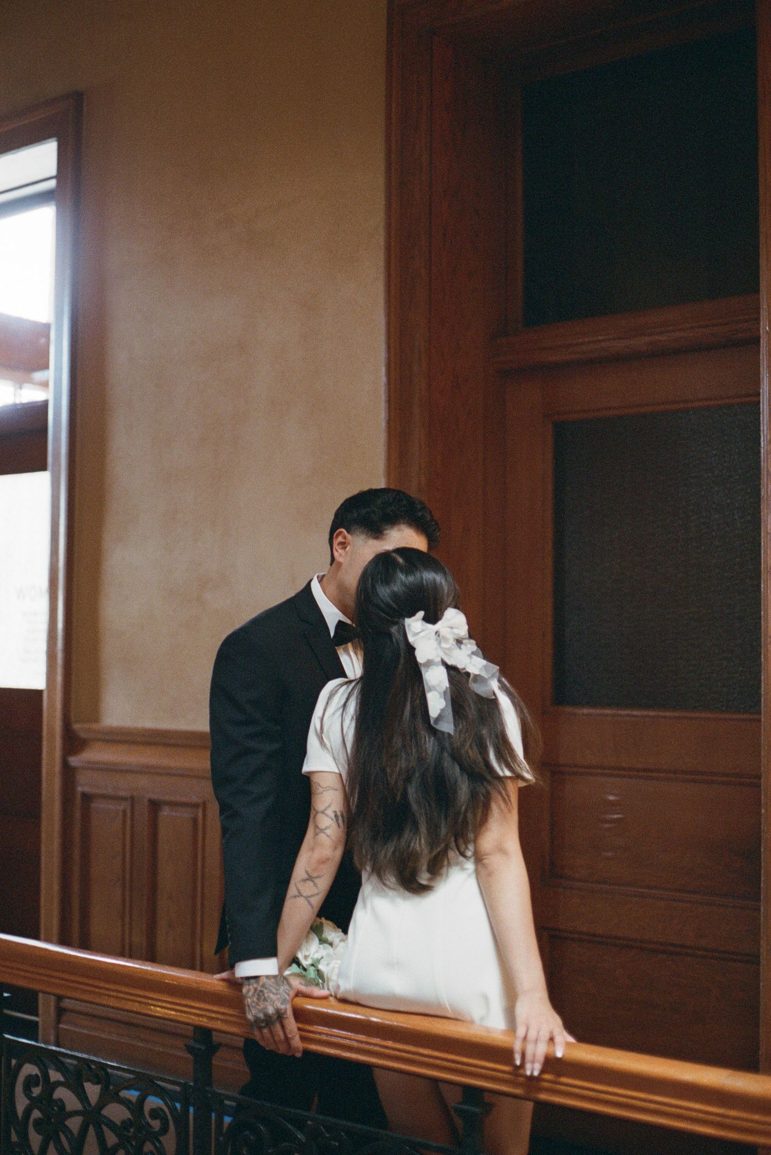 Groom in black tuxedo kisses bride with white bow in her hair on an indoor wooden staircase.