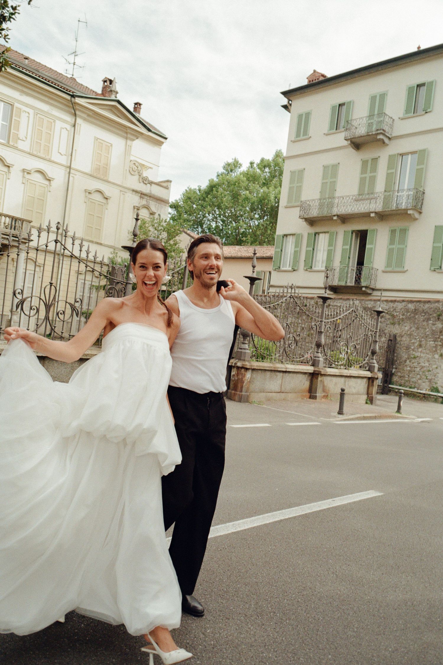 Happy bride and groom laugh while walking on Italian street with historic buildings behind.