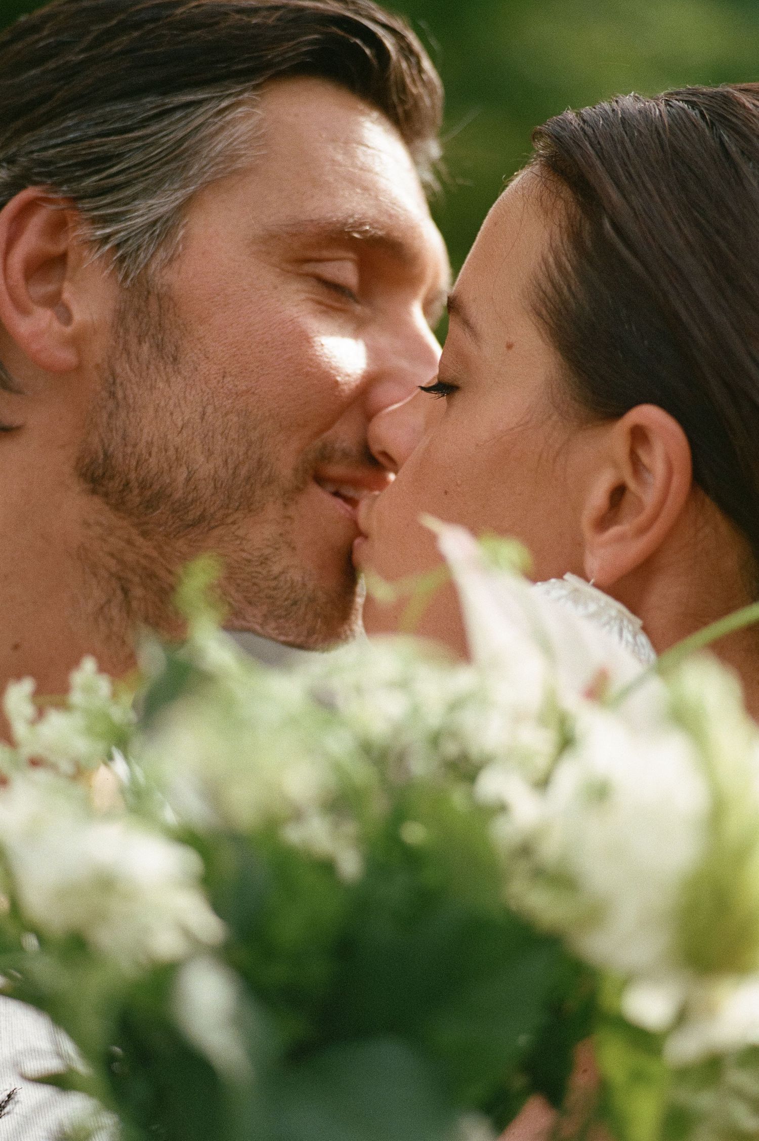 Bride and groom sharing a tender kiss surrounded by a lush white floral bouquet outdoors.