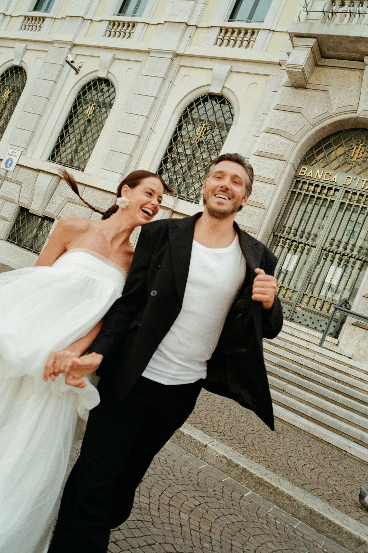 Laughing bride and groom walking outside a Banca d'Italia building in Italy on their wedding day.