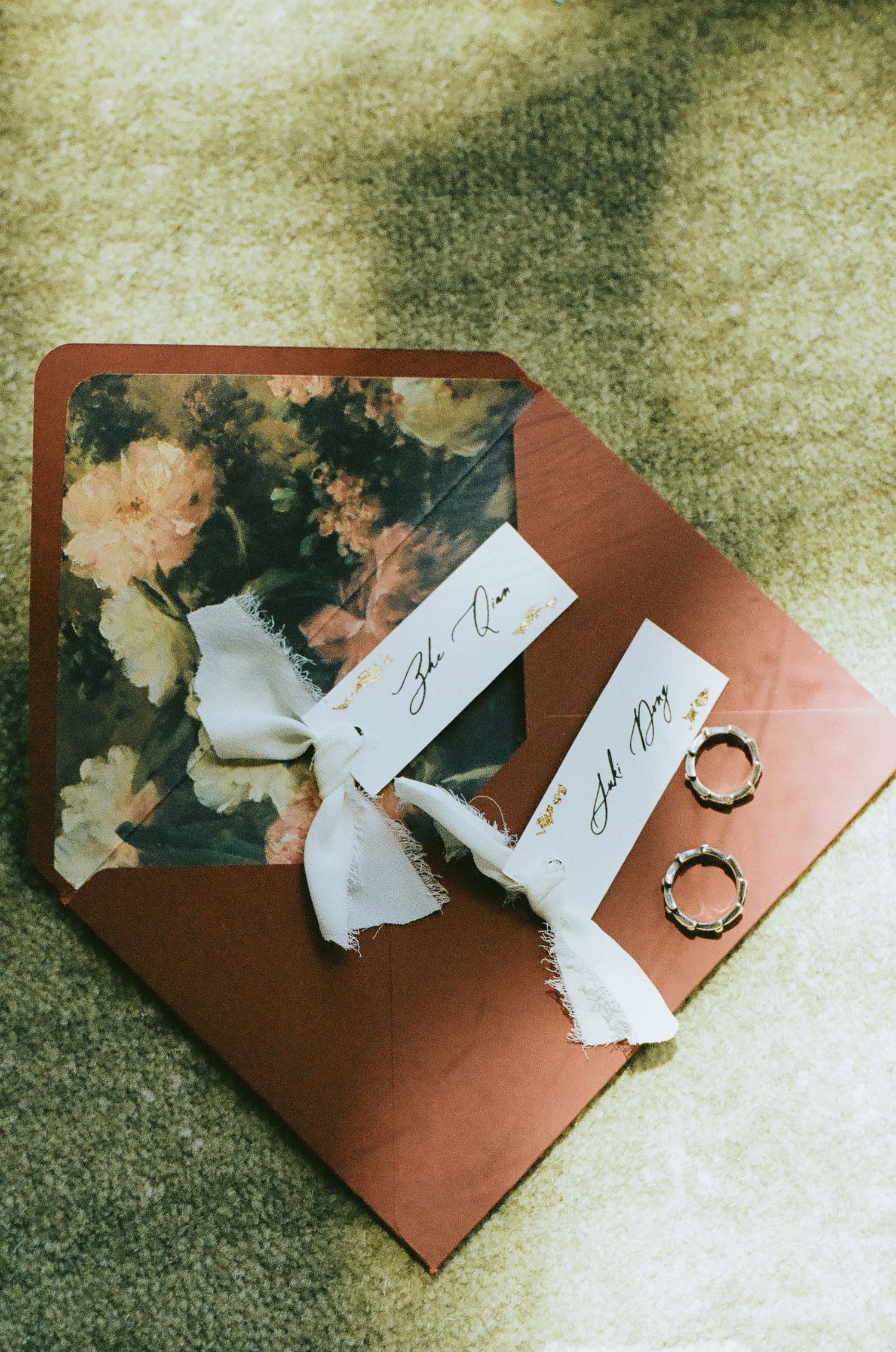 Wedding flat lay with floral envelope, calligraphy name cards, silk ribbon, and gold bamboo rings on green surface.