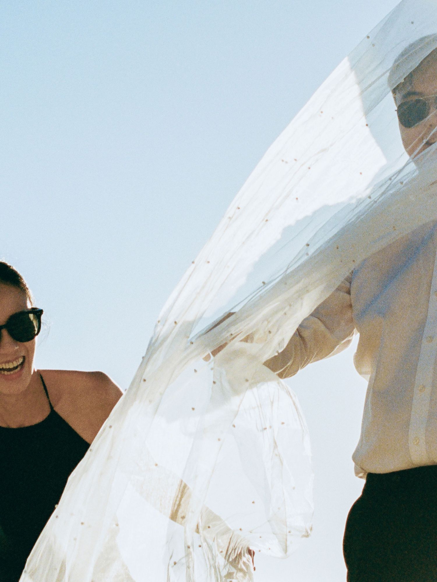 Bride and companion laughing while holding flowing white veil against clear blue sky.