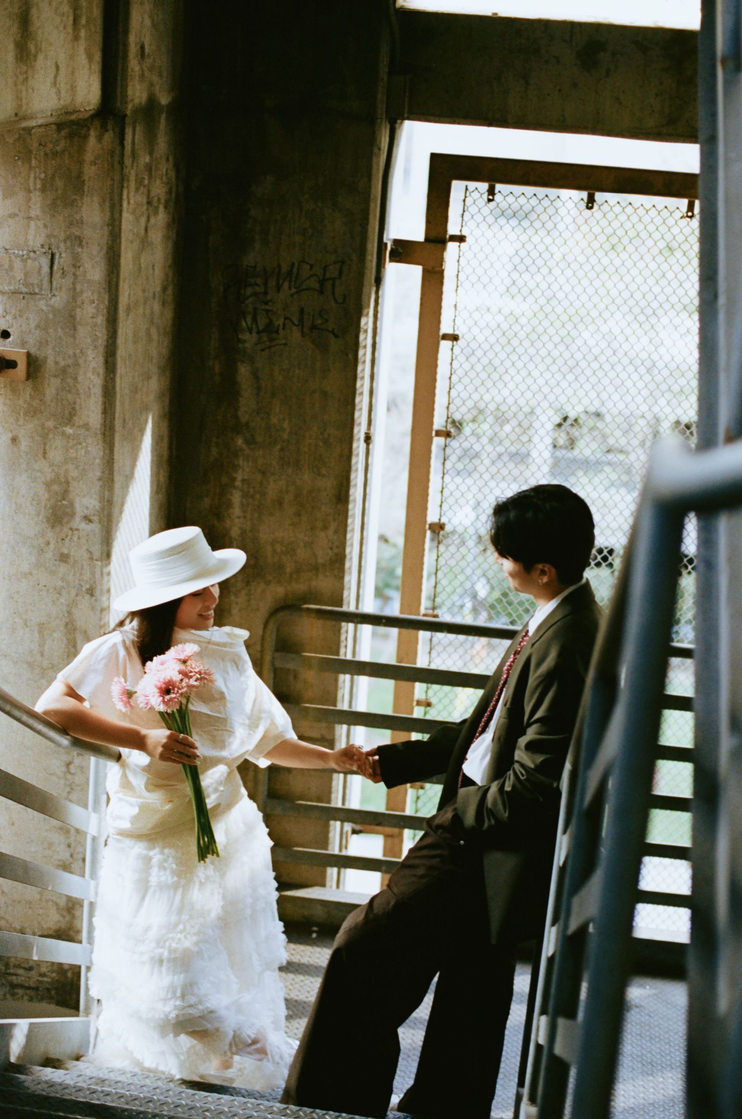 Bride and groom hold hands on a staircase, bride in white dress and hat carrying pink flowers.