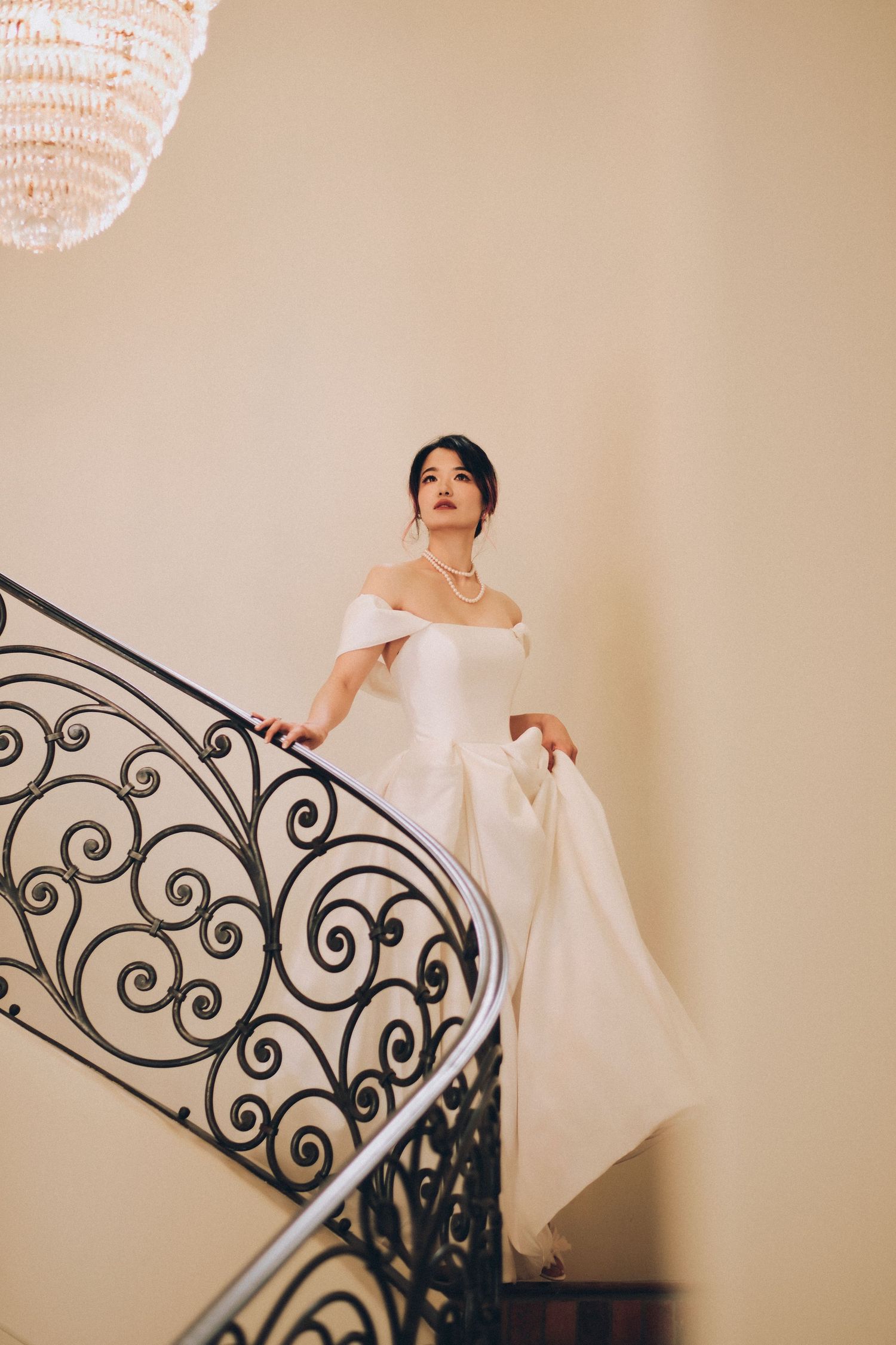 Elegant bride in off-shoulder white gown descending ornate spiral staircase with chandelier.