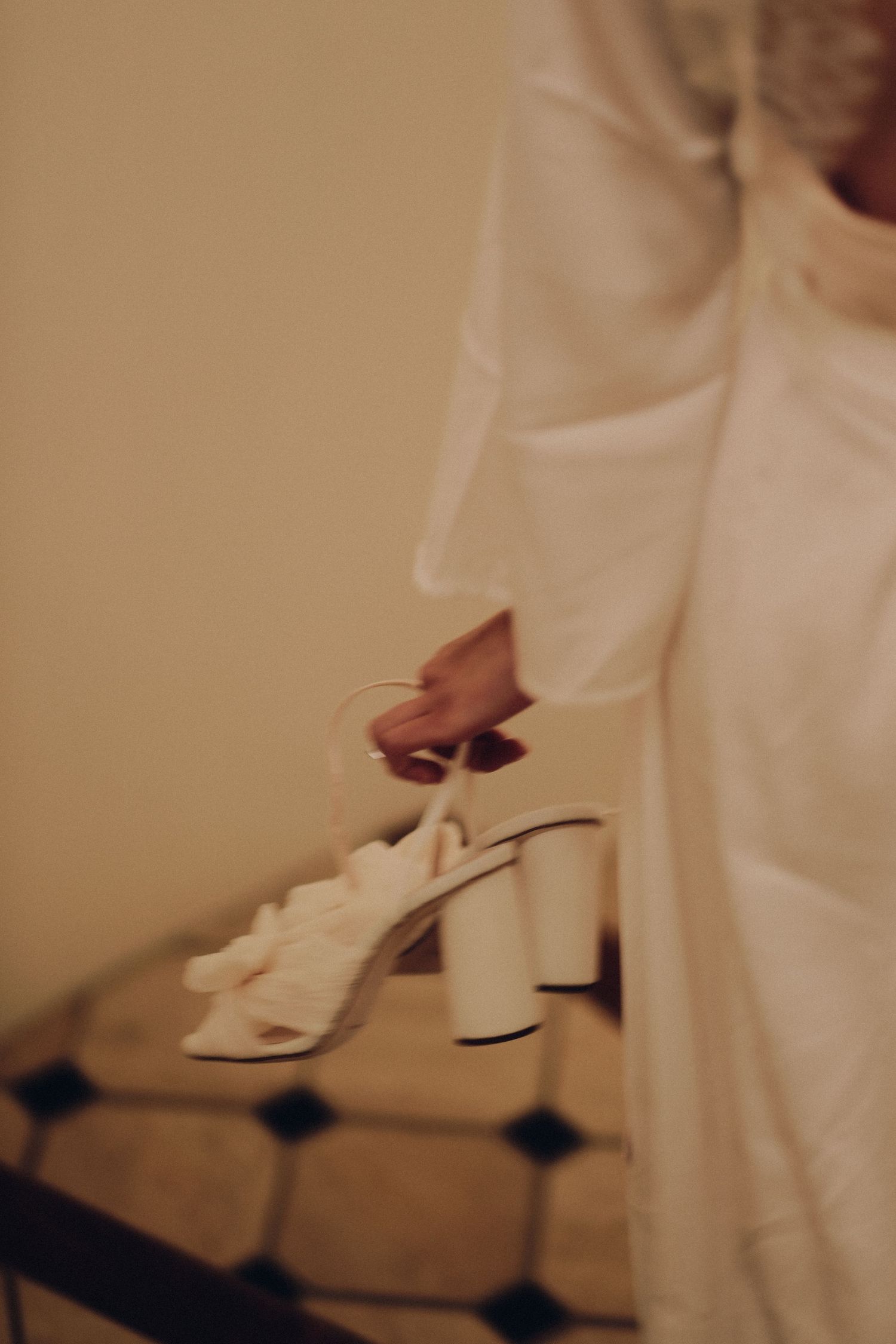 Bride in white wedding dress holding elegant white block heel sandals with floral detail on staircase.