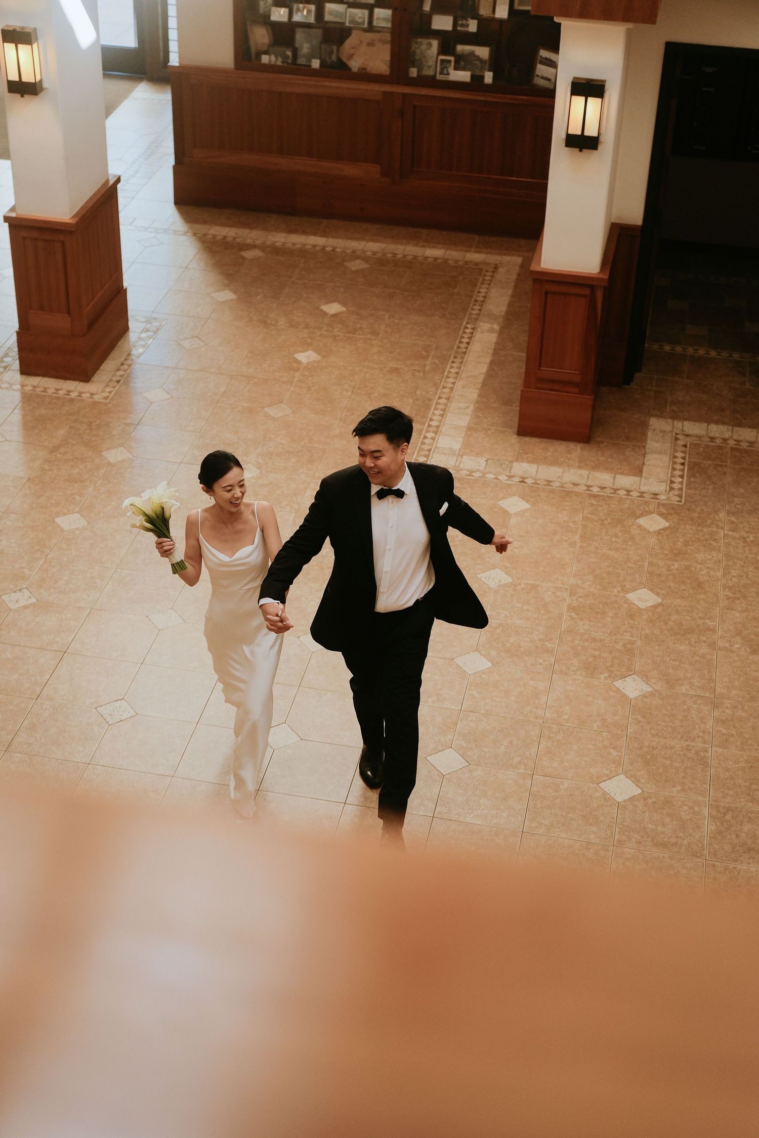 Overhead view of Asian couple in wedding attire walking through hotel lobby, bride holding white calla lily bouquet.