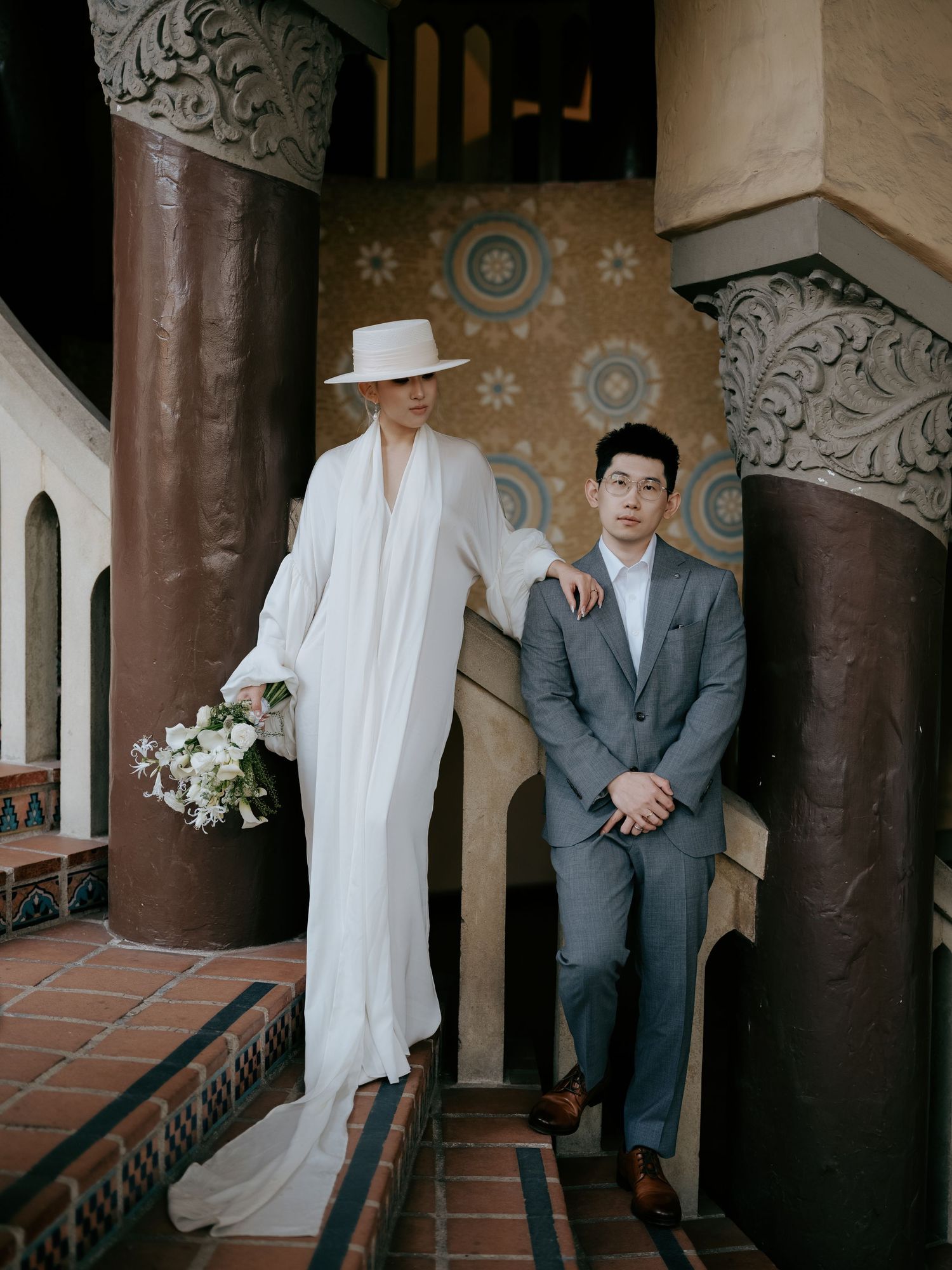 Elegant bride in white cape and wide-brim hat poses with groom beside ornate decorative columns.
