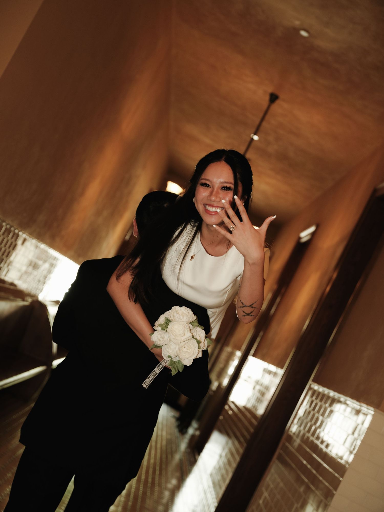 Smiling bride holds white bouquet and shows wedding ring in golden elevator mirror reflection.