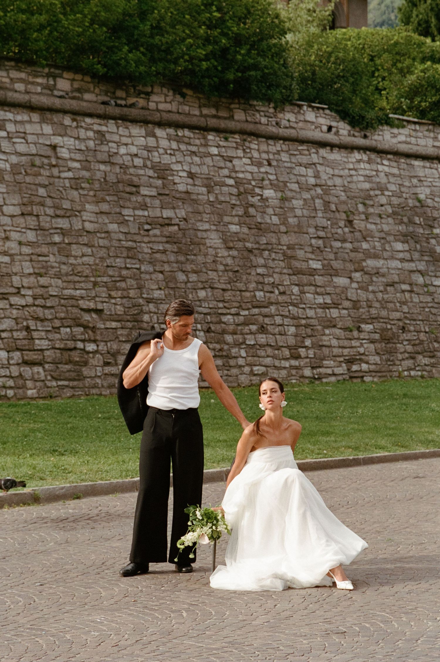 Bride in white strapless gown crouches holding bouquet while groom adjusts jacket on cobblestone path.