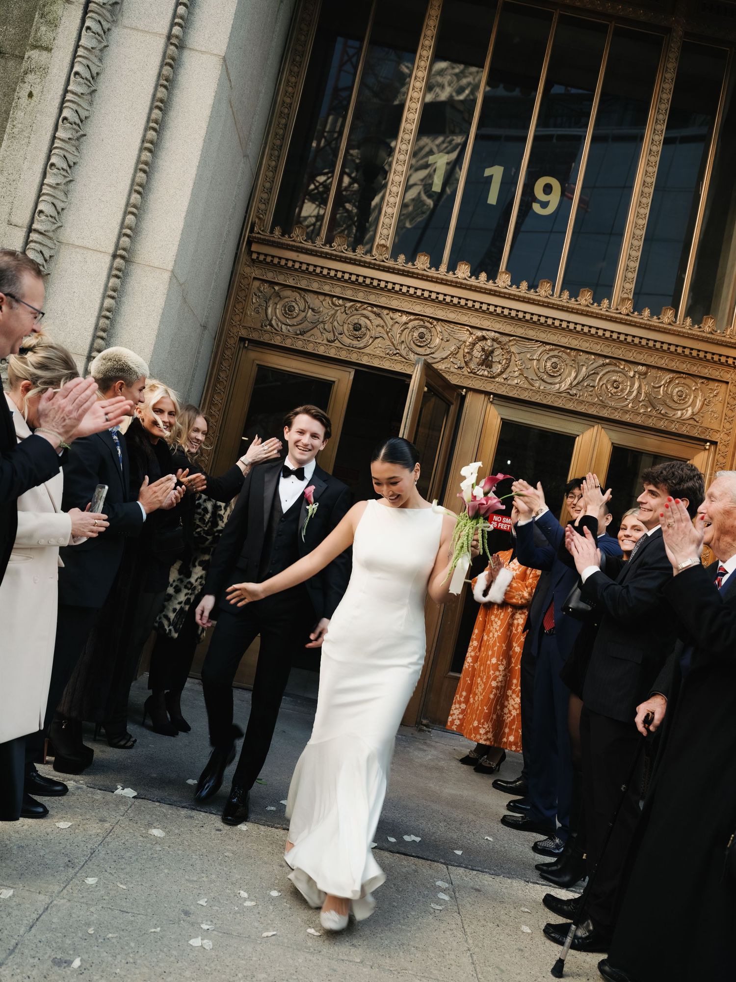 Newlyweds exit grand Art Deco building as guests celebrate with applause and confetti in New York.