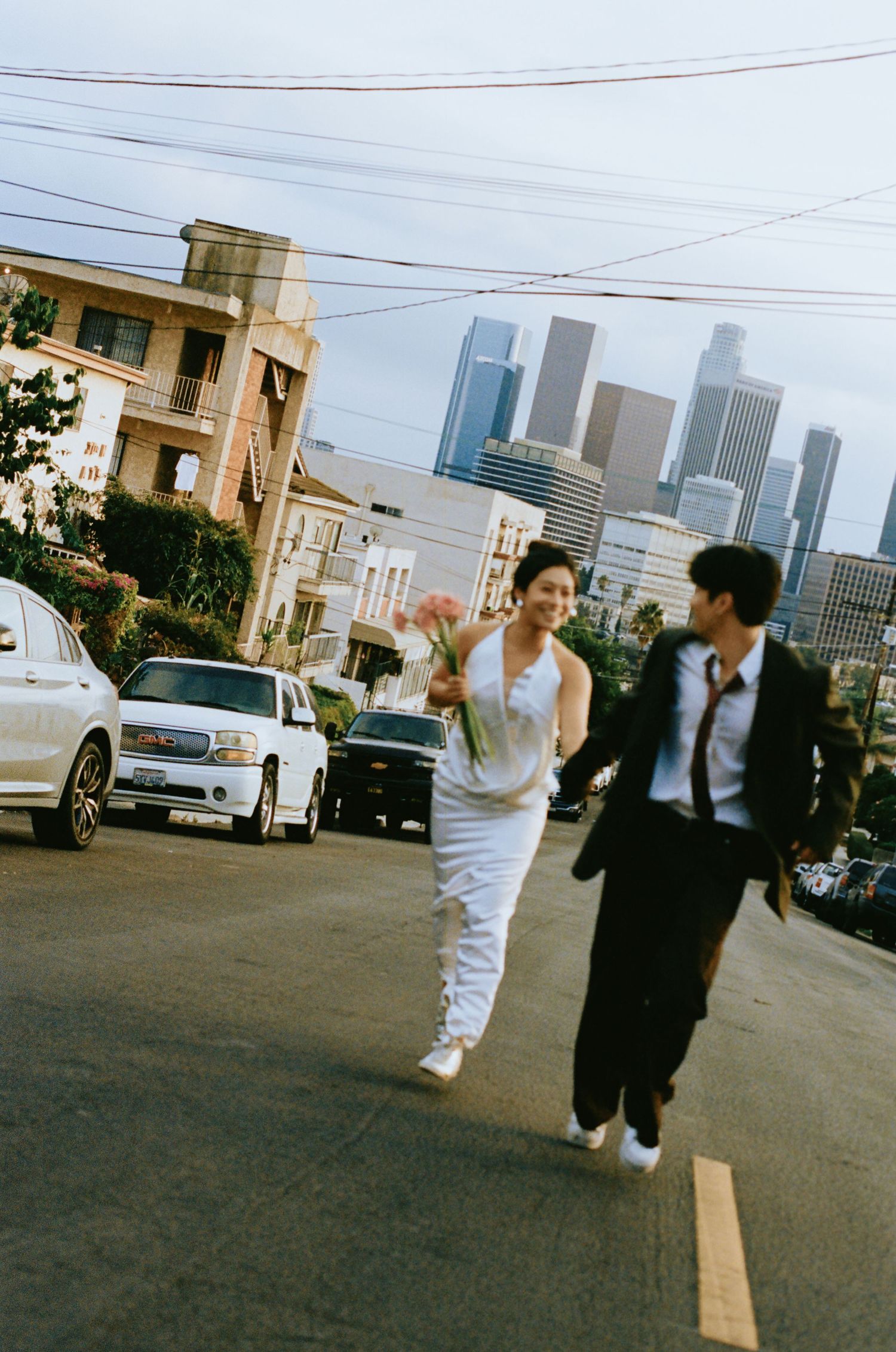 Bride and groom run hand-in-hand across a street with Los Angeles skyline in the background.