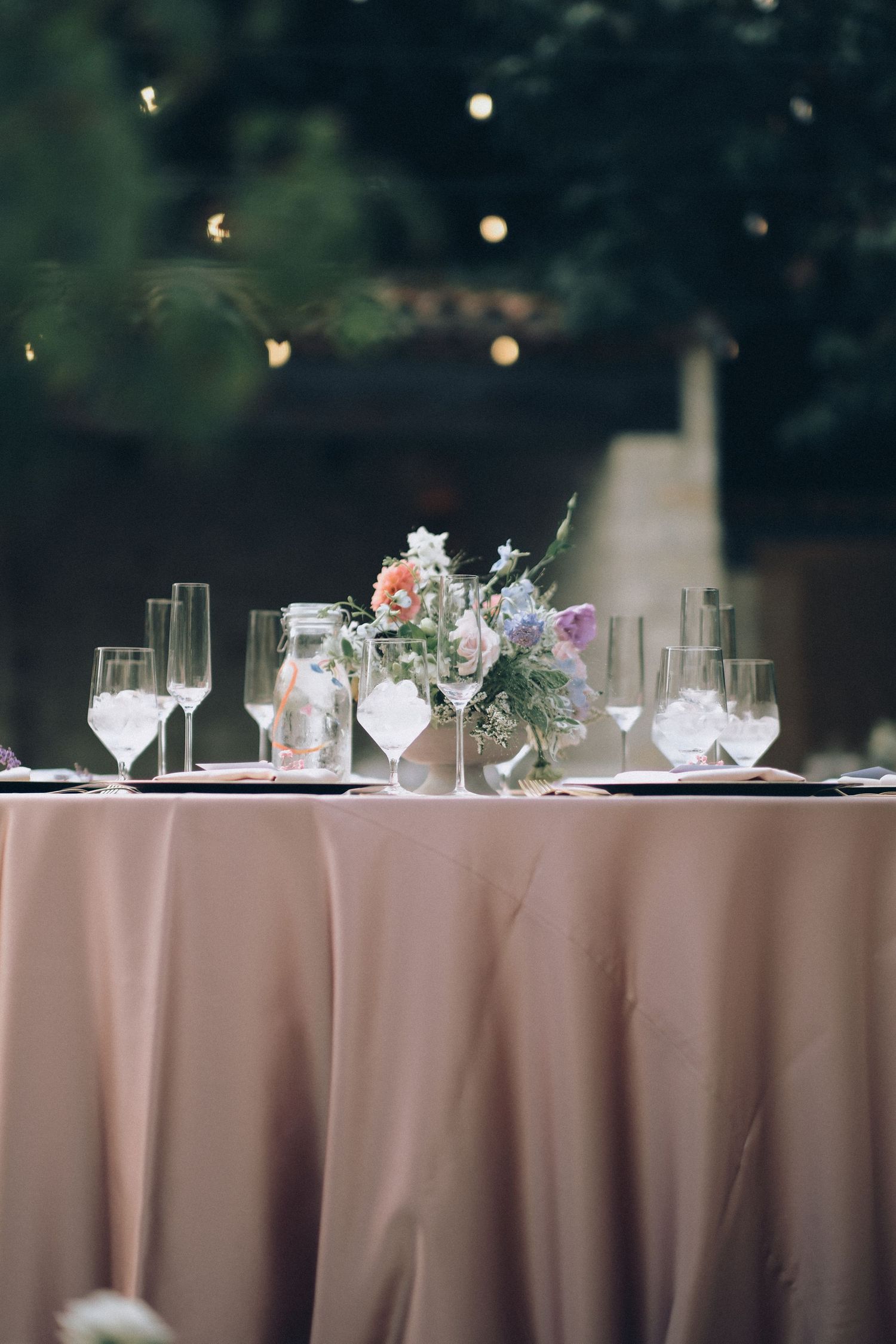 Elegant wedding reception table with blush pink linen, crystal glassware, and a small floral centerpiece under string lights.