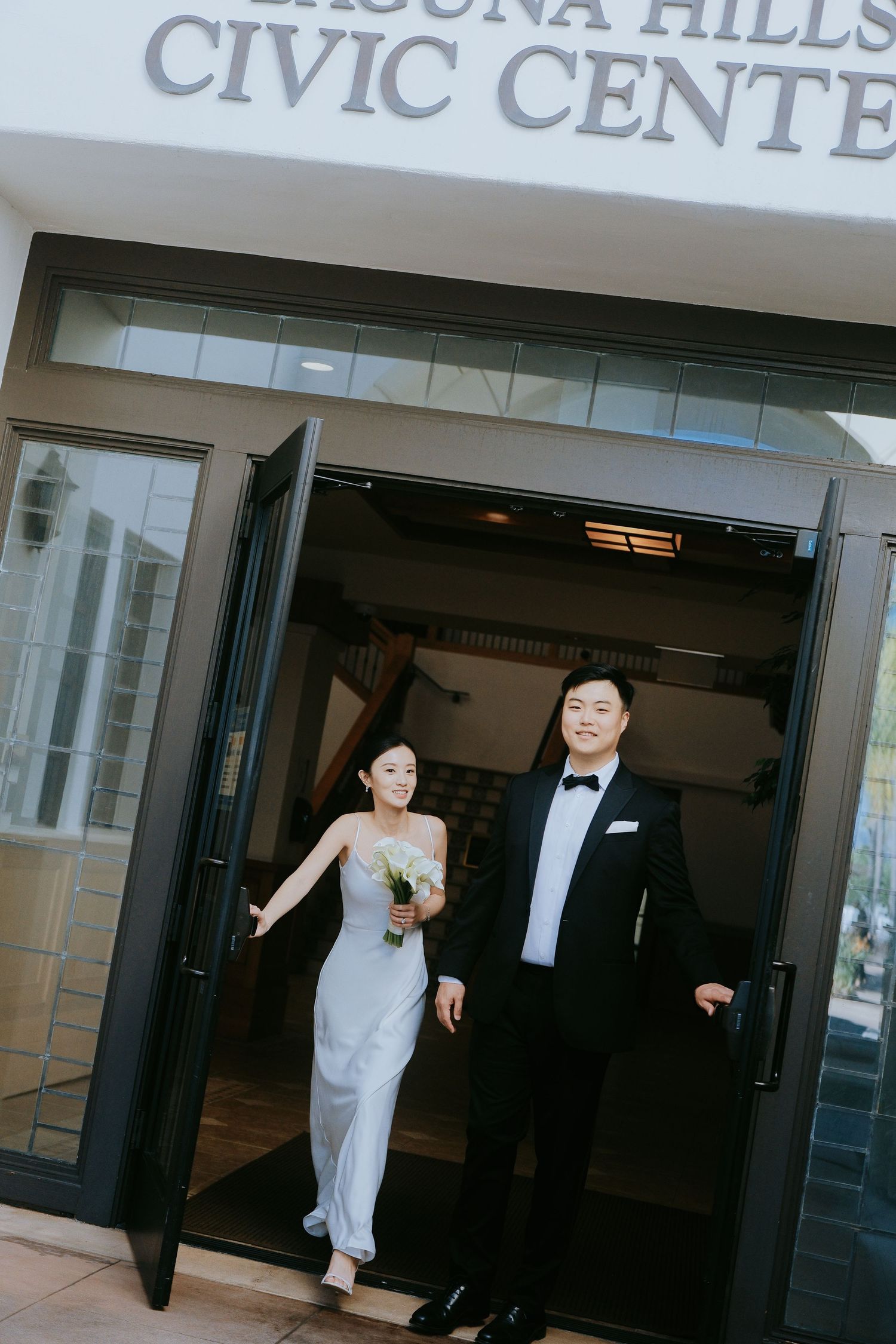Asian bride and groom exiting Laguna Hills Civic Center, bride in white gown holding calla lily bouquet.